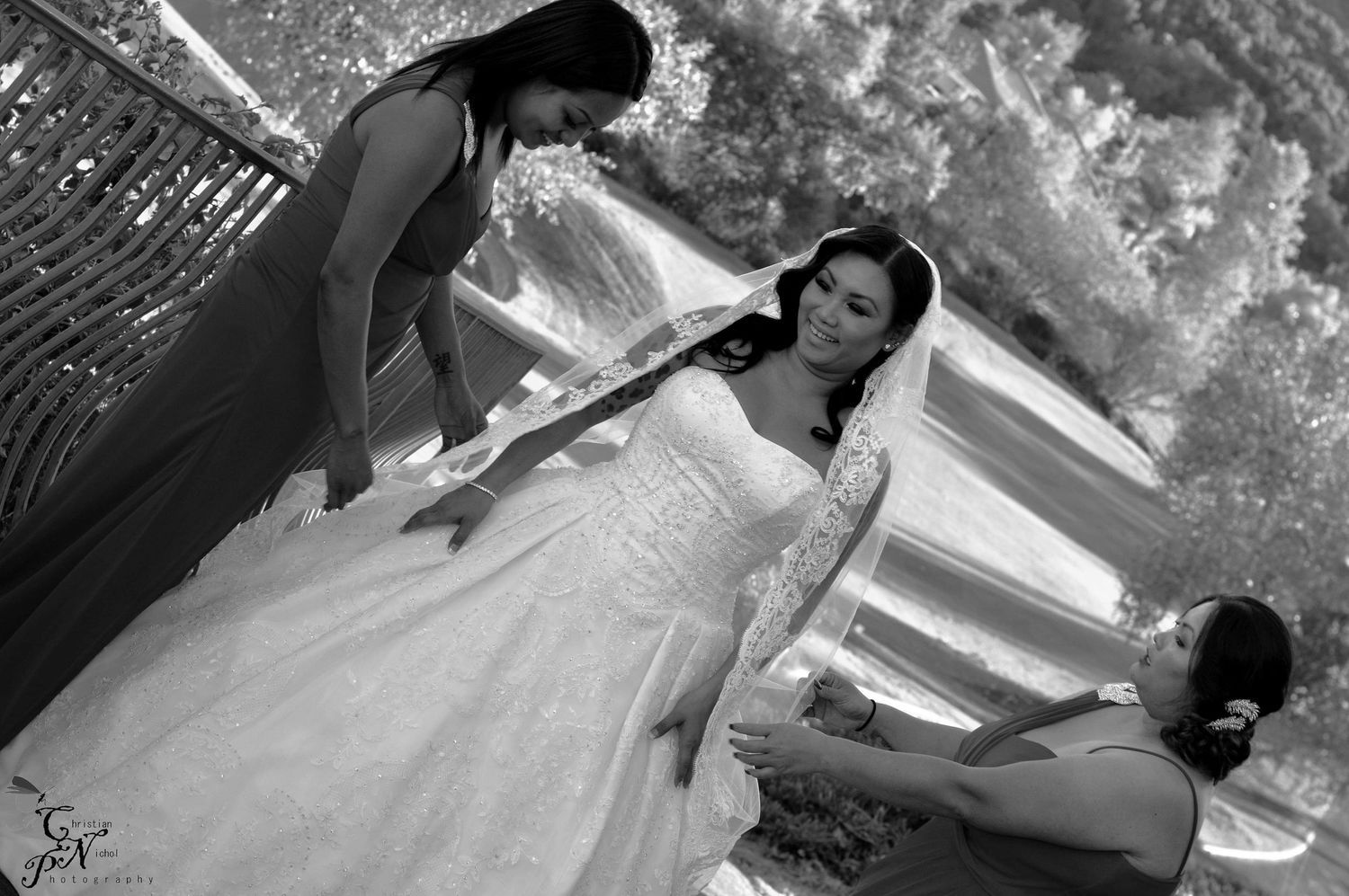 Black and white photo of a bride in a white gown being assisted by two bridesmaids outdoors.