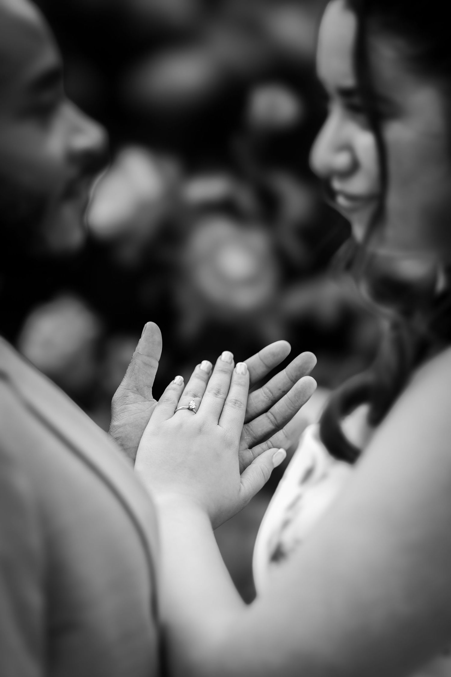 Black and white photo of couple holding hands, showing engagement ring, faces softly blurred in romantic bokeh background.