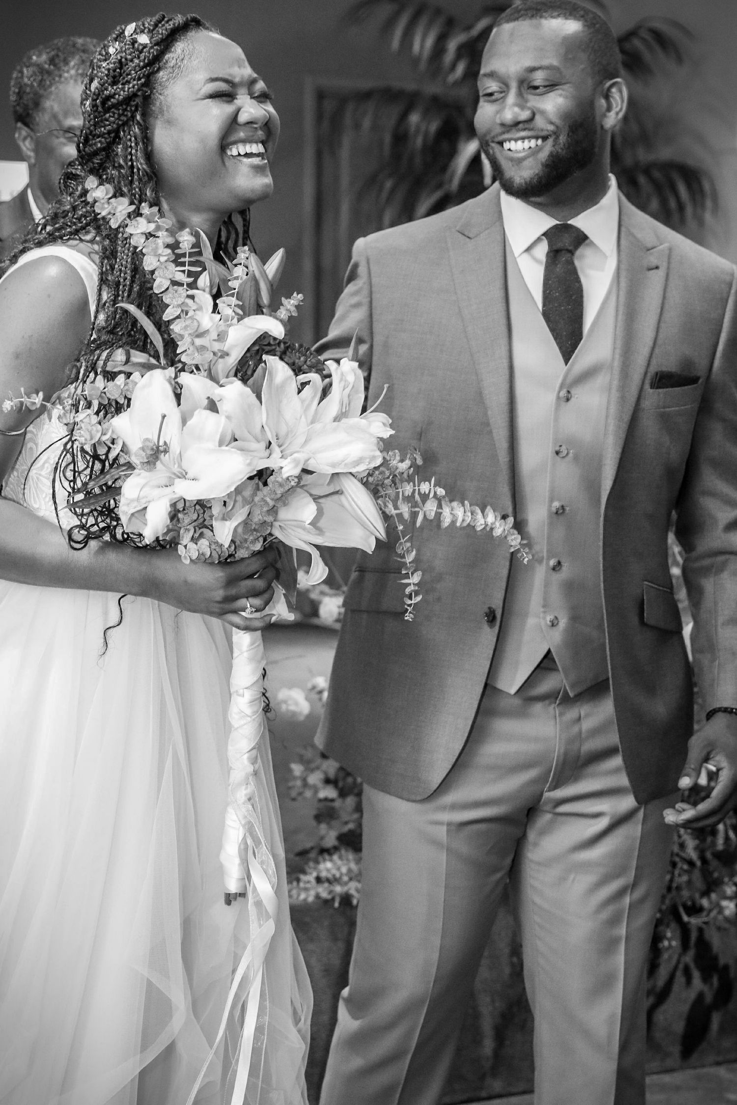Black and white photo of a joyful bride holding a floral bouquet and groom in a suit smiling at their wedding ceremony.