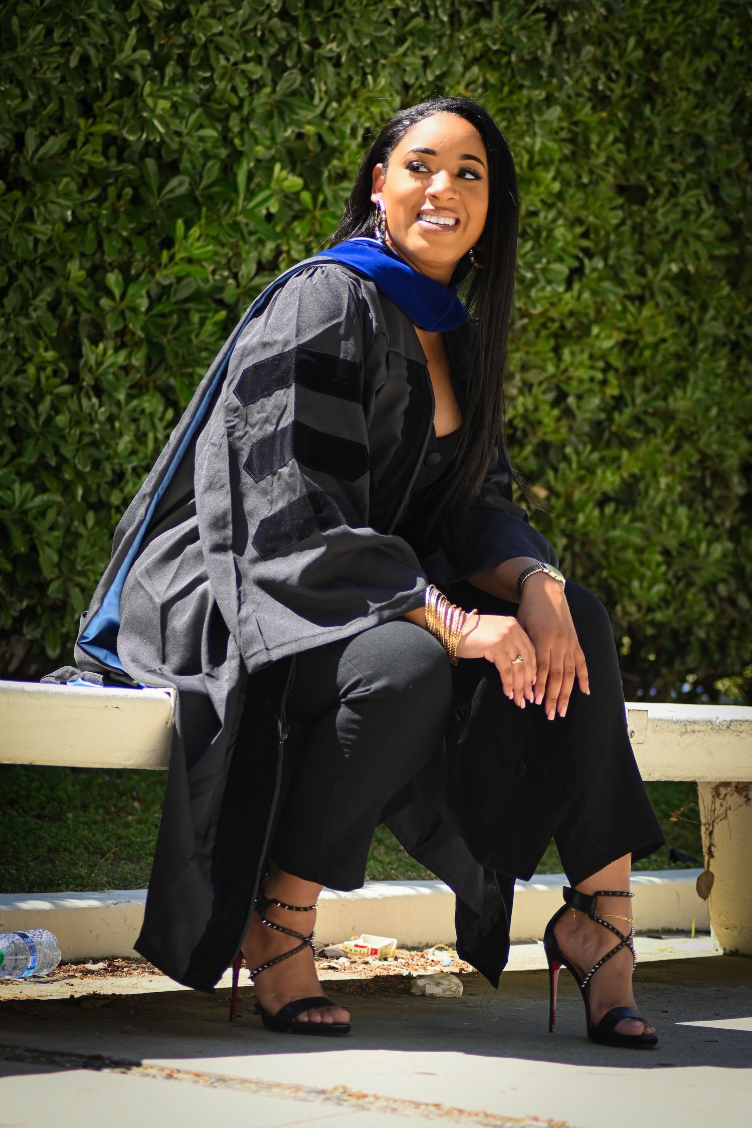 Graduate in cap and gown sits casually on white bench during photo session.