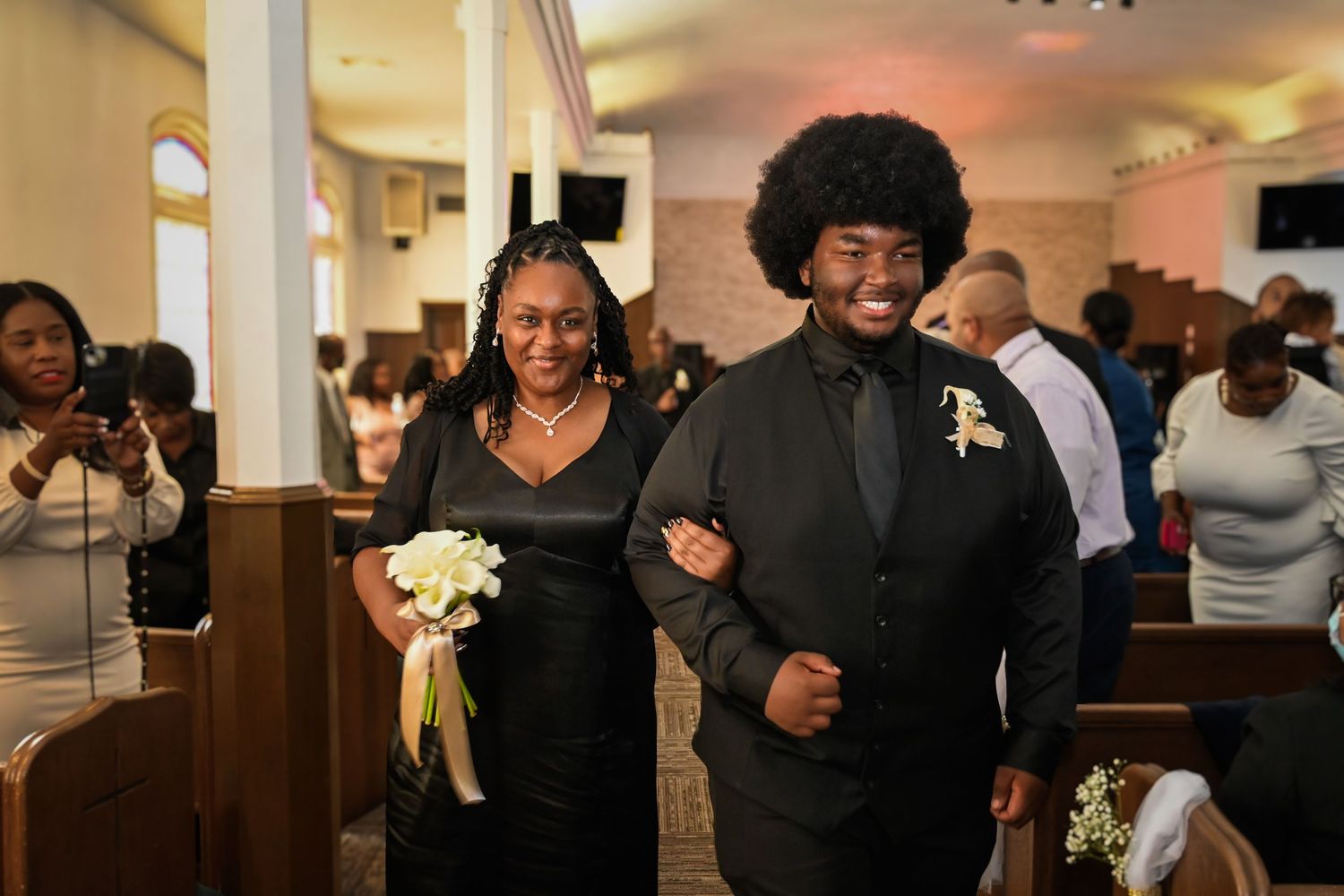 A young man in a black suit escorts a woman in a black dress down a church aisle during a wedding ceremony.