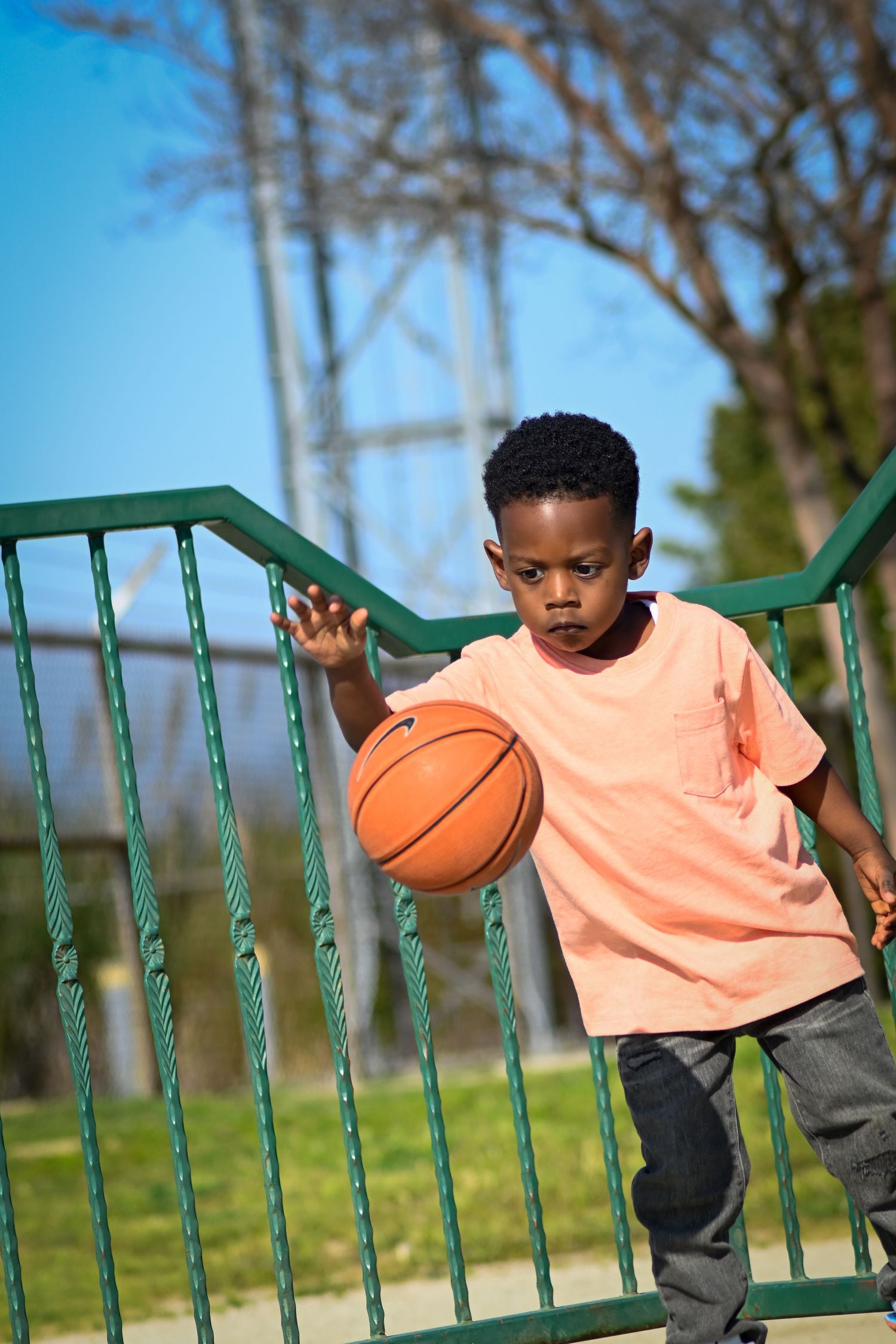 Young Black boy dribbling an orange basketball outdoors near green railings on a sunny day.
