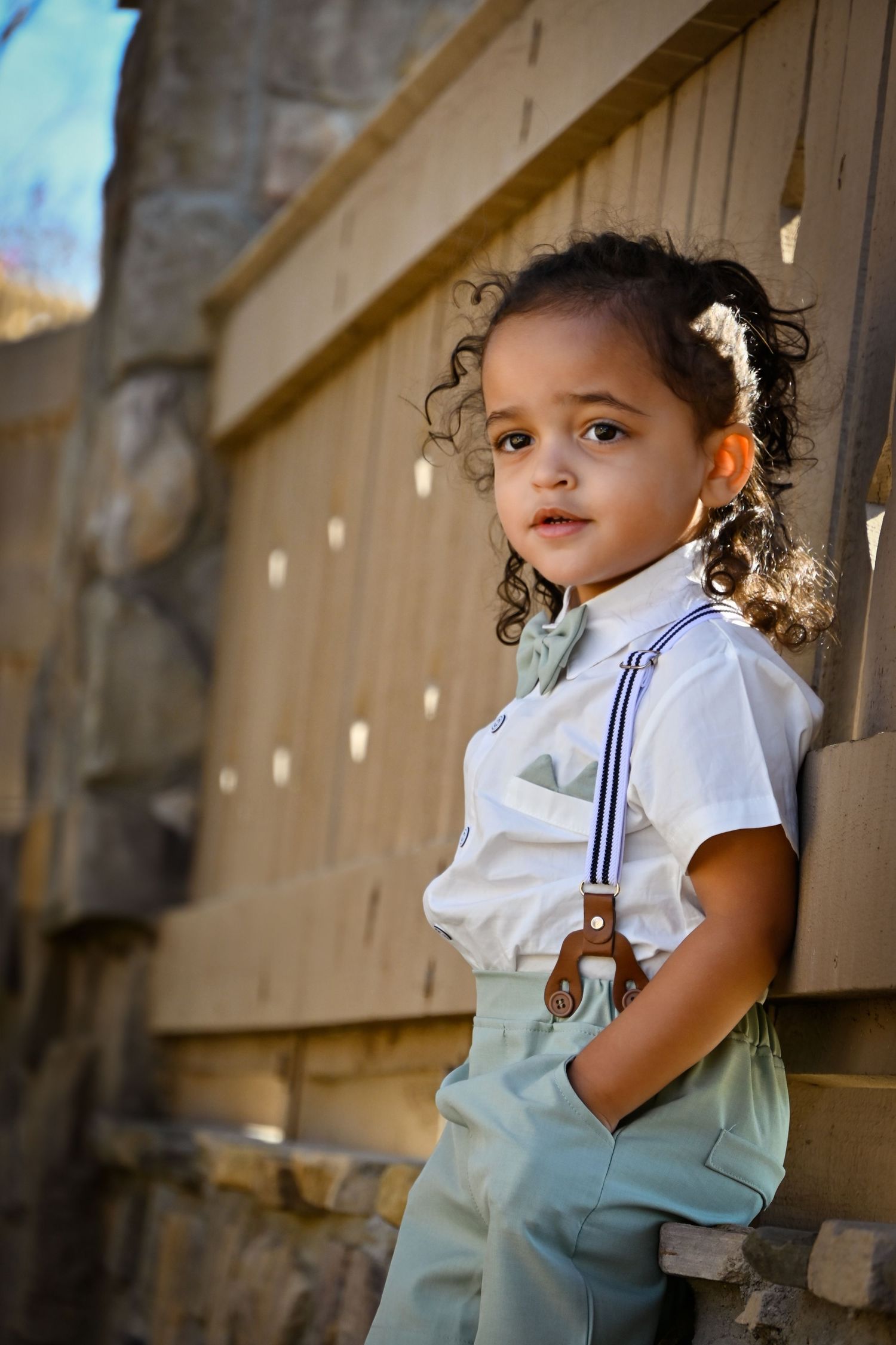 A young child in white shirt with suspenders and mint pants leans against a wooden fence outdoors.