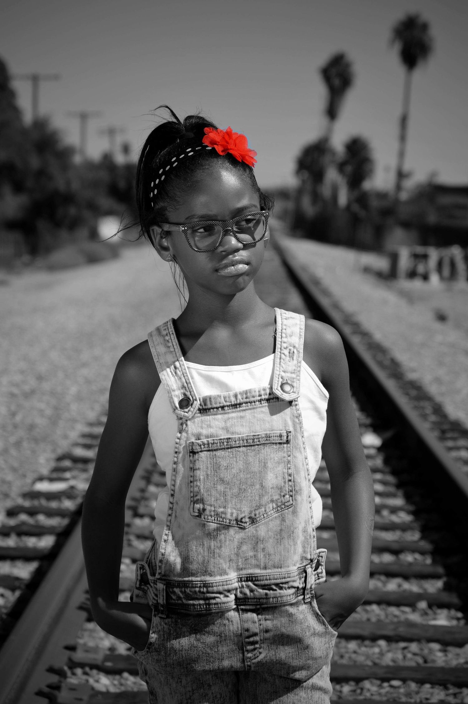 A young Black girl in denim overalls and glasses stands on railroad tracks, with a red flower in her hair.
