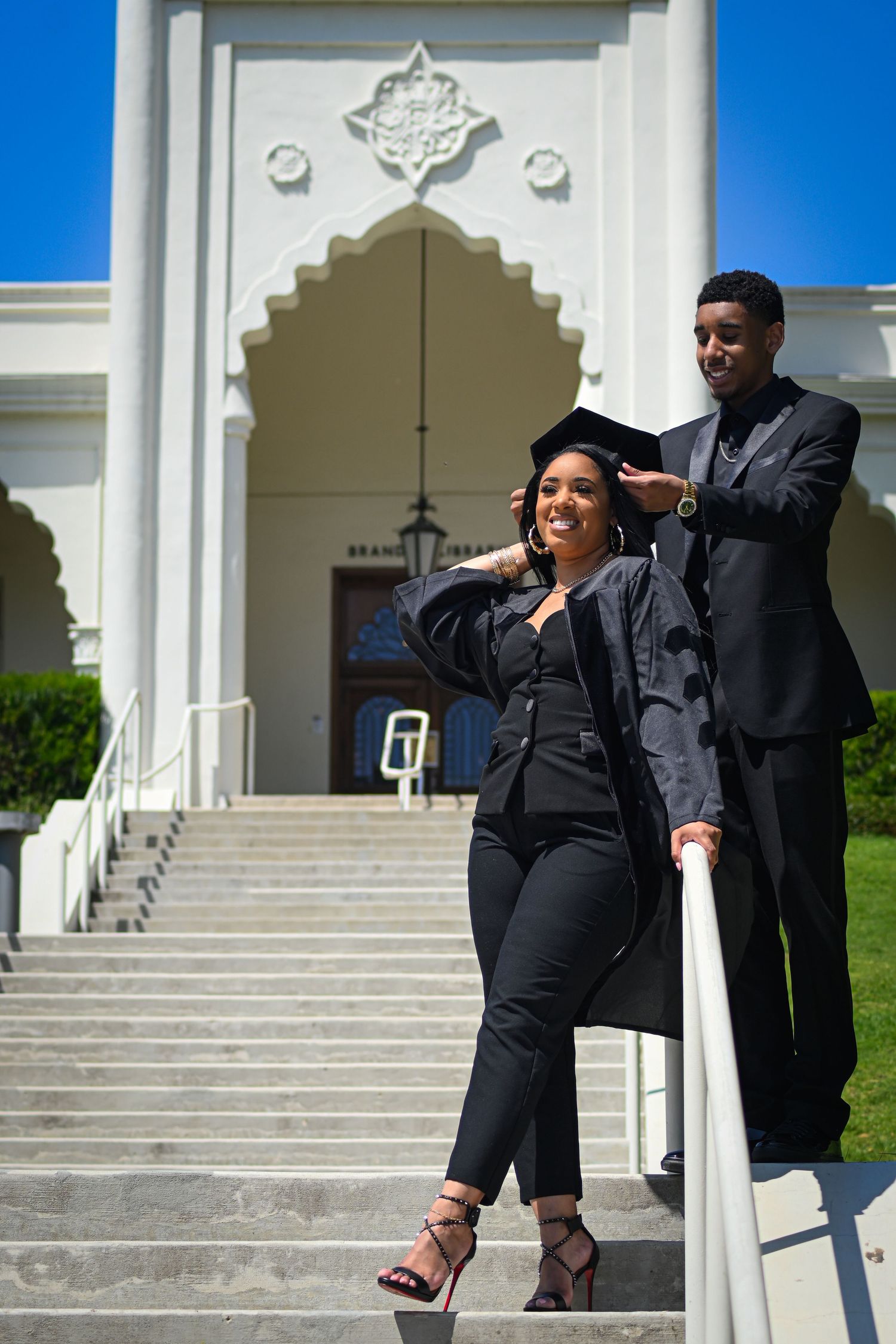 Graduate in black attire poses confidently on steps of ornate building with architectural details.
