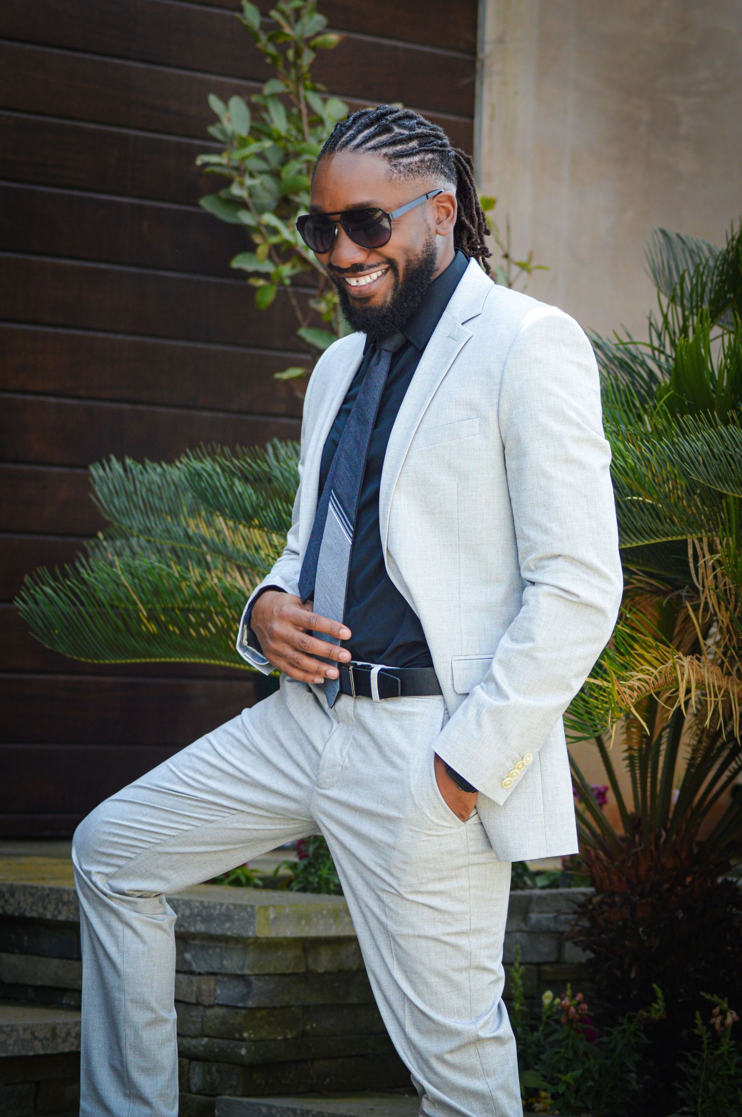 Stylishly dressed person in a white suit and sunglasses poses confidently against a backdrop of tropical plants.