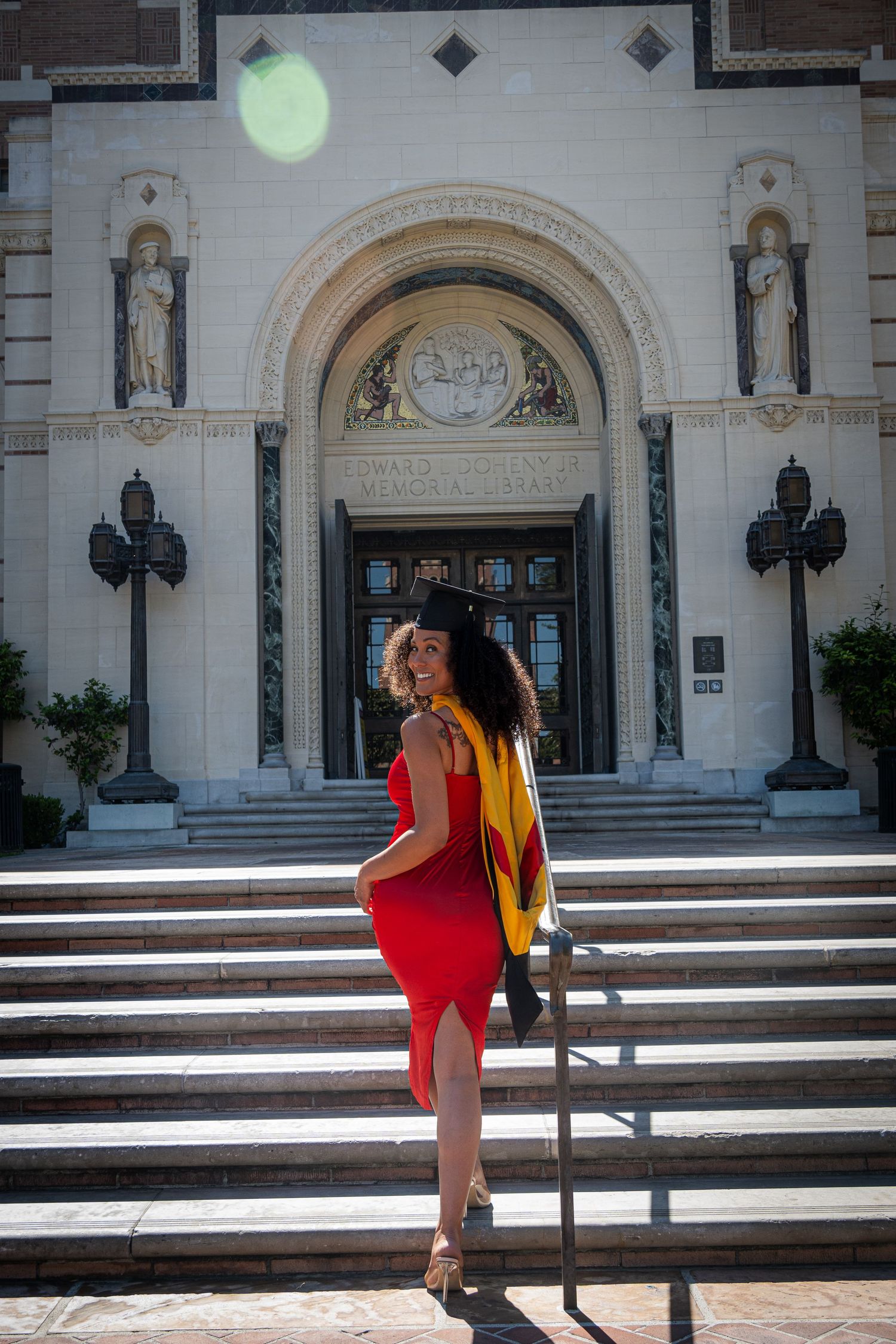 Graduate ascending white stone steps in red dress with gold graduation stole at historic building entrance.