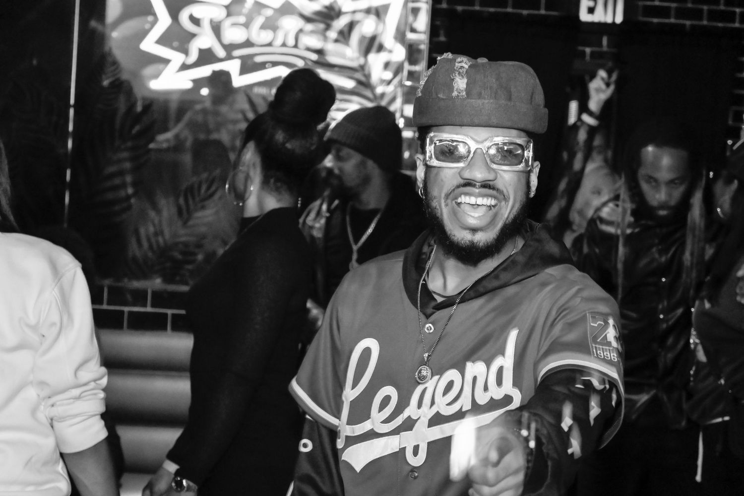 A person wearing glasses and a baseball jersey smiles at a nightclub with brick walls in the background.
