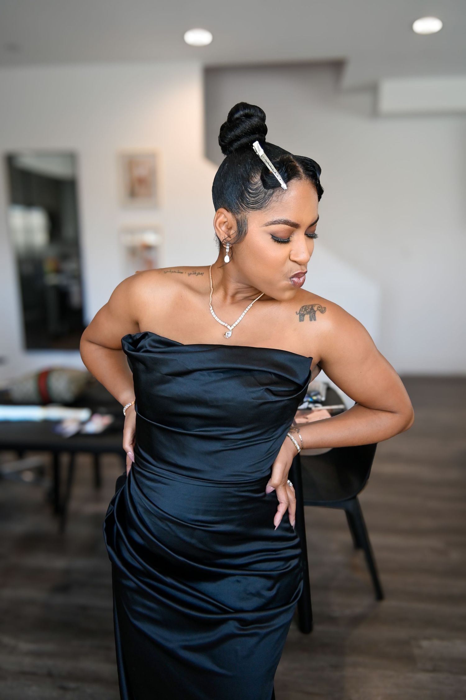 Woman in elegant black strapless satin gown with updo hairstyle and silver jewelry poses indoors.