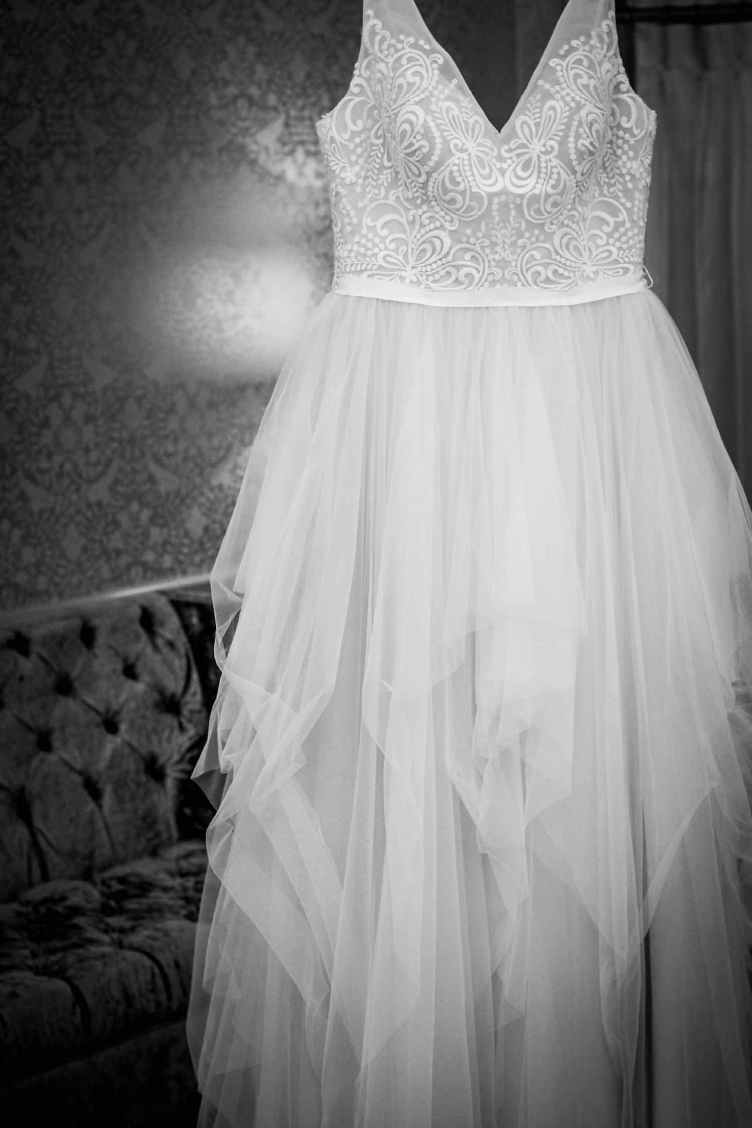 Black and white photo of an elegant lace bodice wedding dress with flowing tulle skirt hanging in a decorated room.