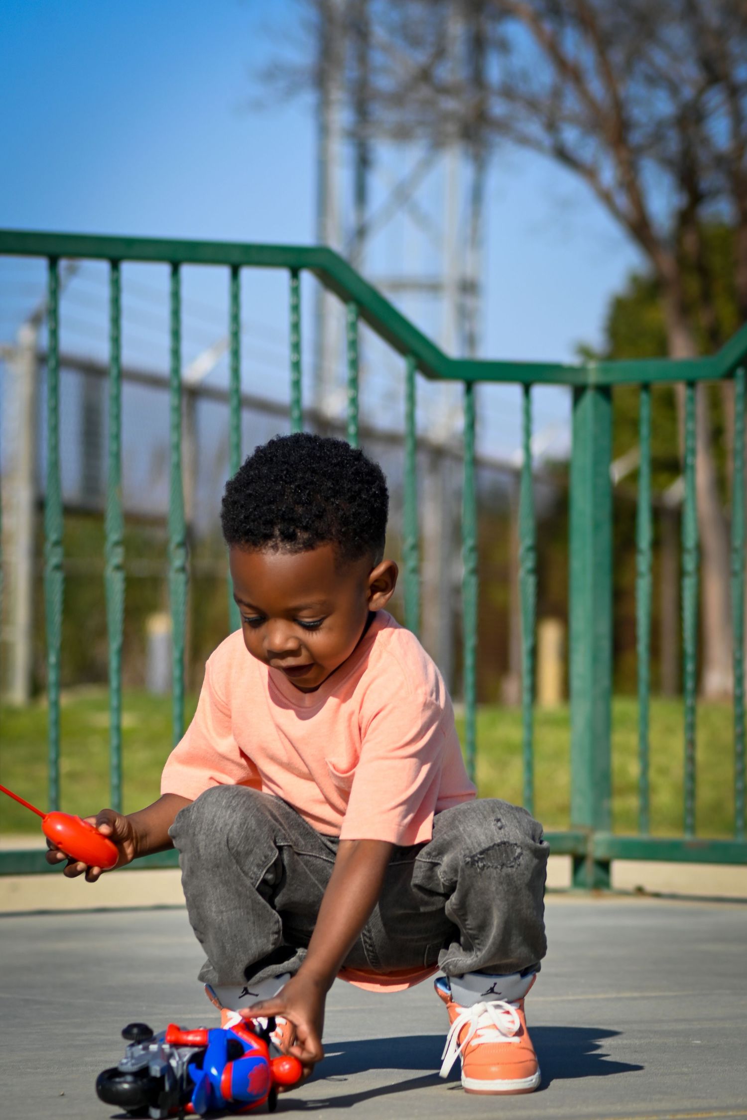 Young Black boy playing with remote control toy car on playground, wearing pink shirt and jeans on sunny day.
