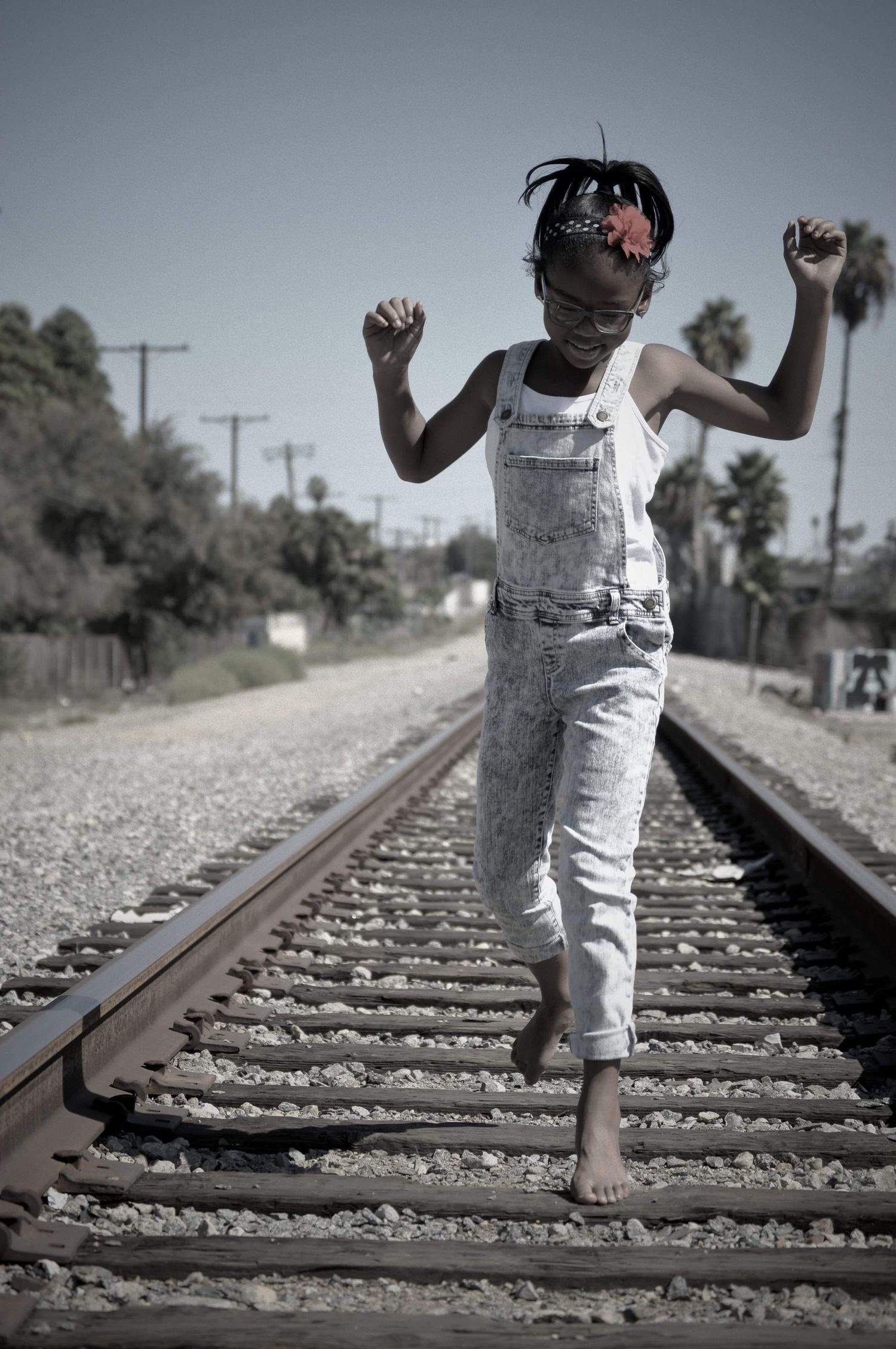 A young girl in white overalls balances barefoot on railroad tracks, arms raised, smiling joyfully on a sunny day.