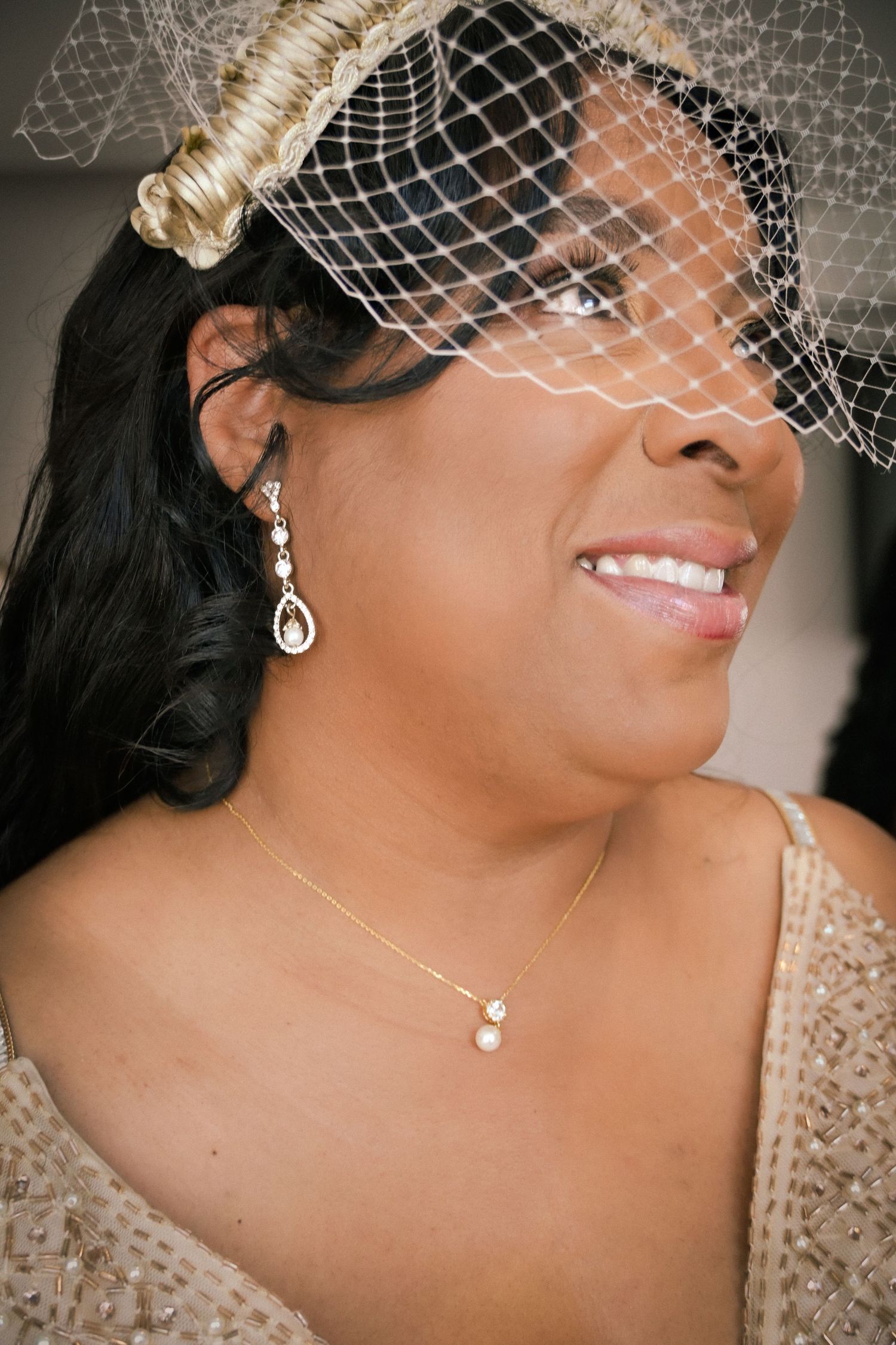 Smiling bride wearing a birdcage veil, crystal drop earrings, and pearl necklace on her wedding day.
