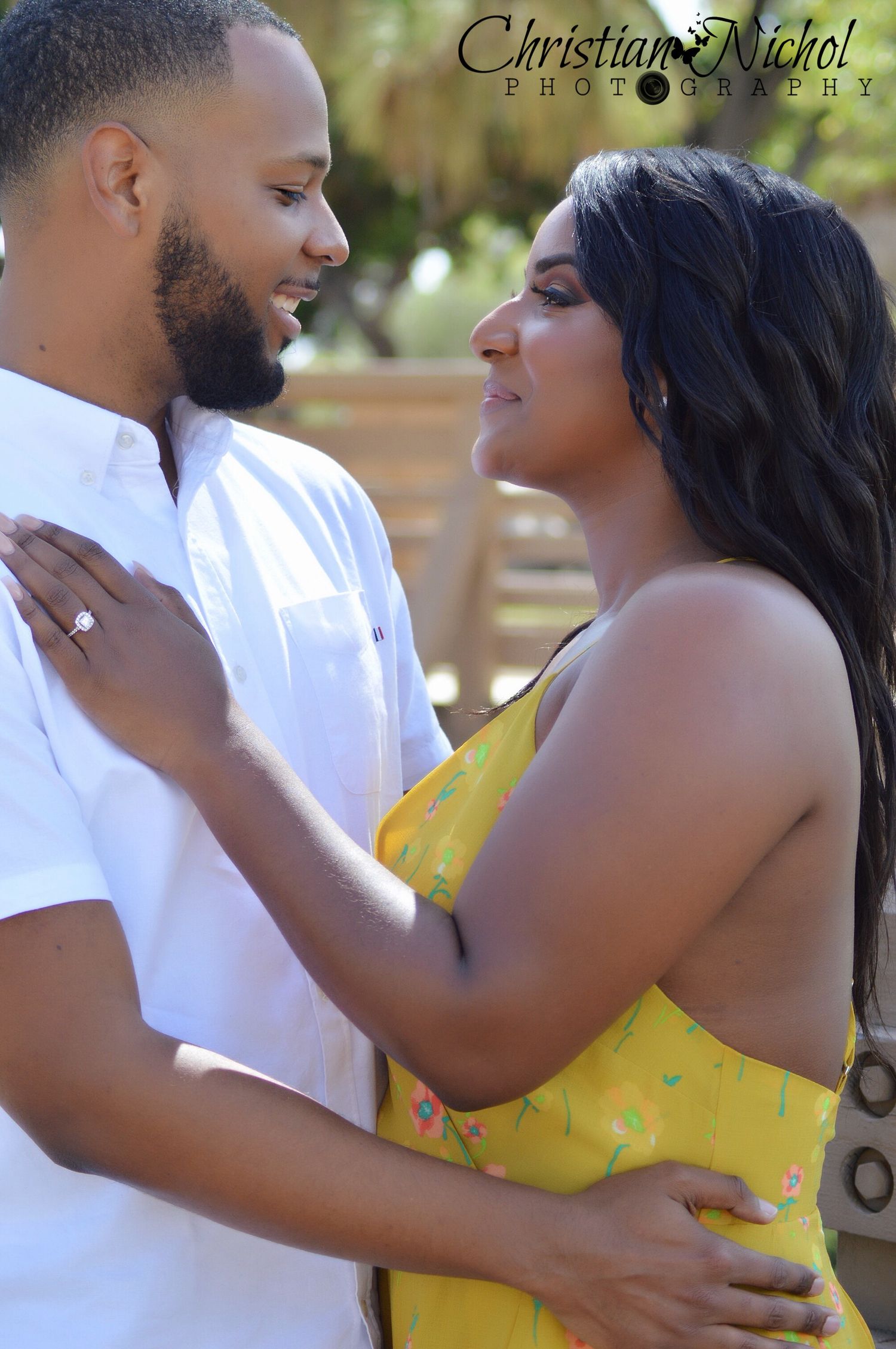 Couple in love gazing at each other, woman in yellow floral dress with engagement ring, man in white shirt, outdoor photo.