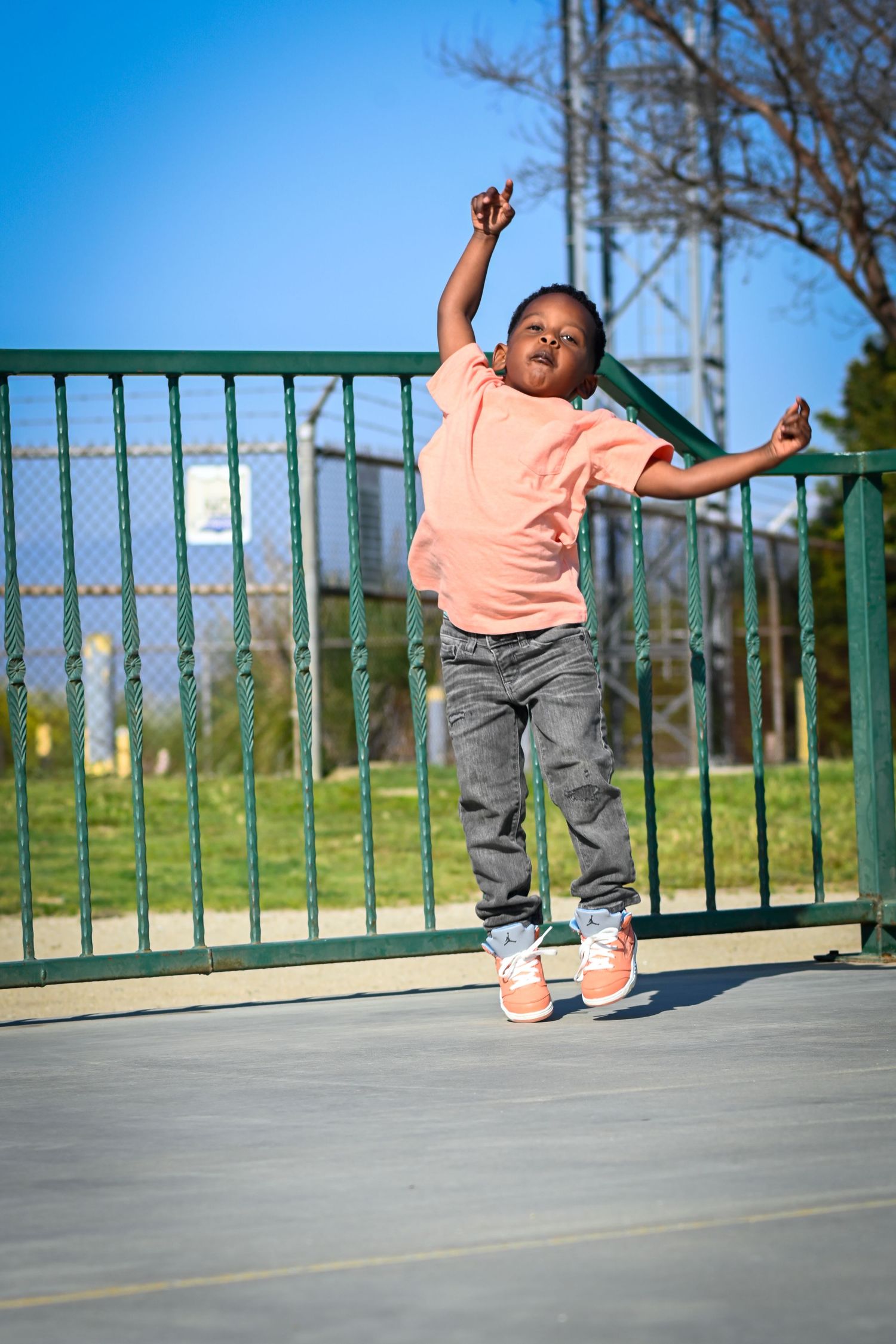 Young Black boy jumping joyfully at a park playground, wearing a pink shirt, jeans, and orange sneakers on a sunny day.