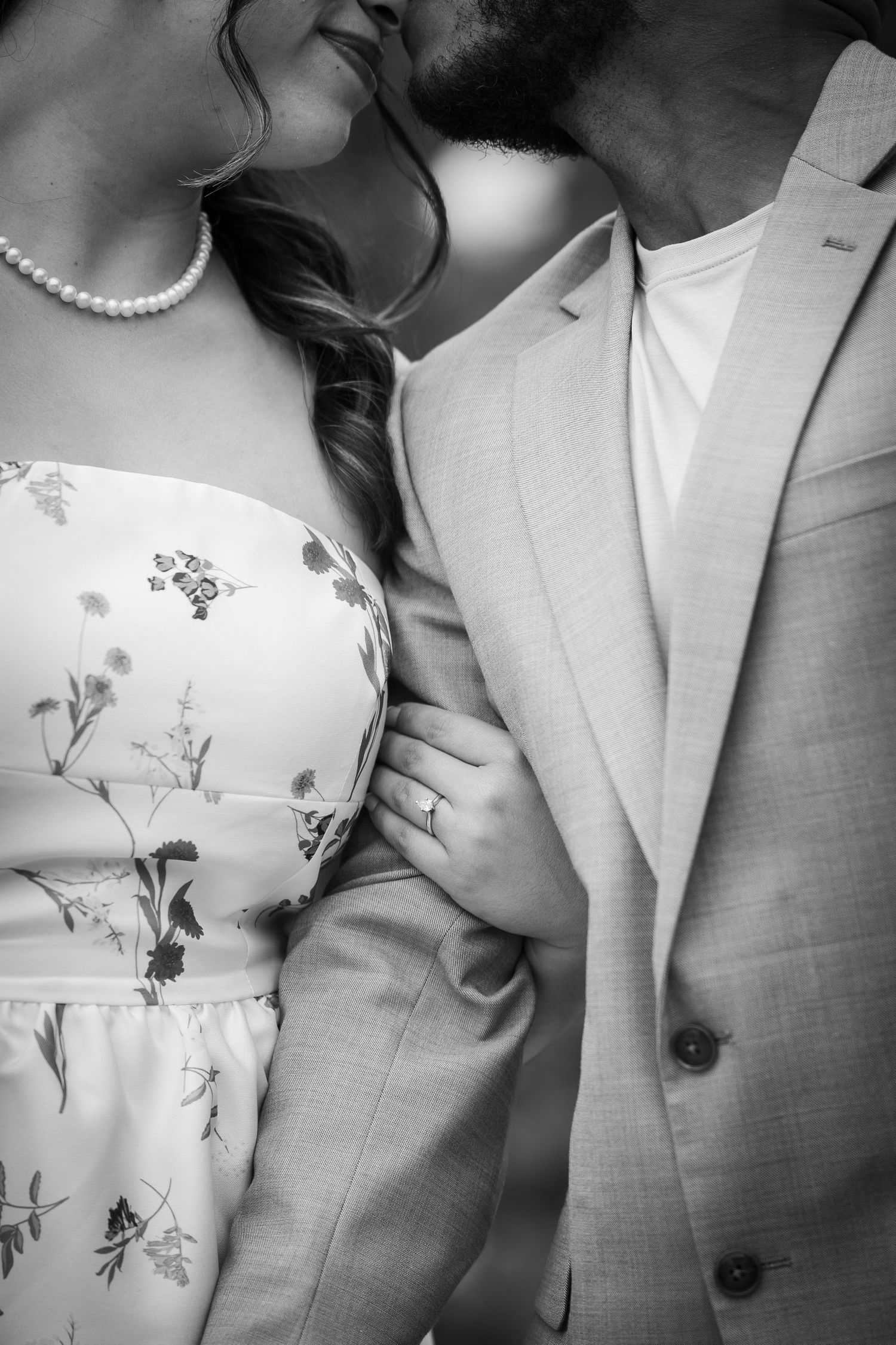 Black and white photo of an engaged couple embracing, woman wearing floral dress with pearl necklace and engagement ring.