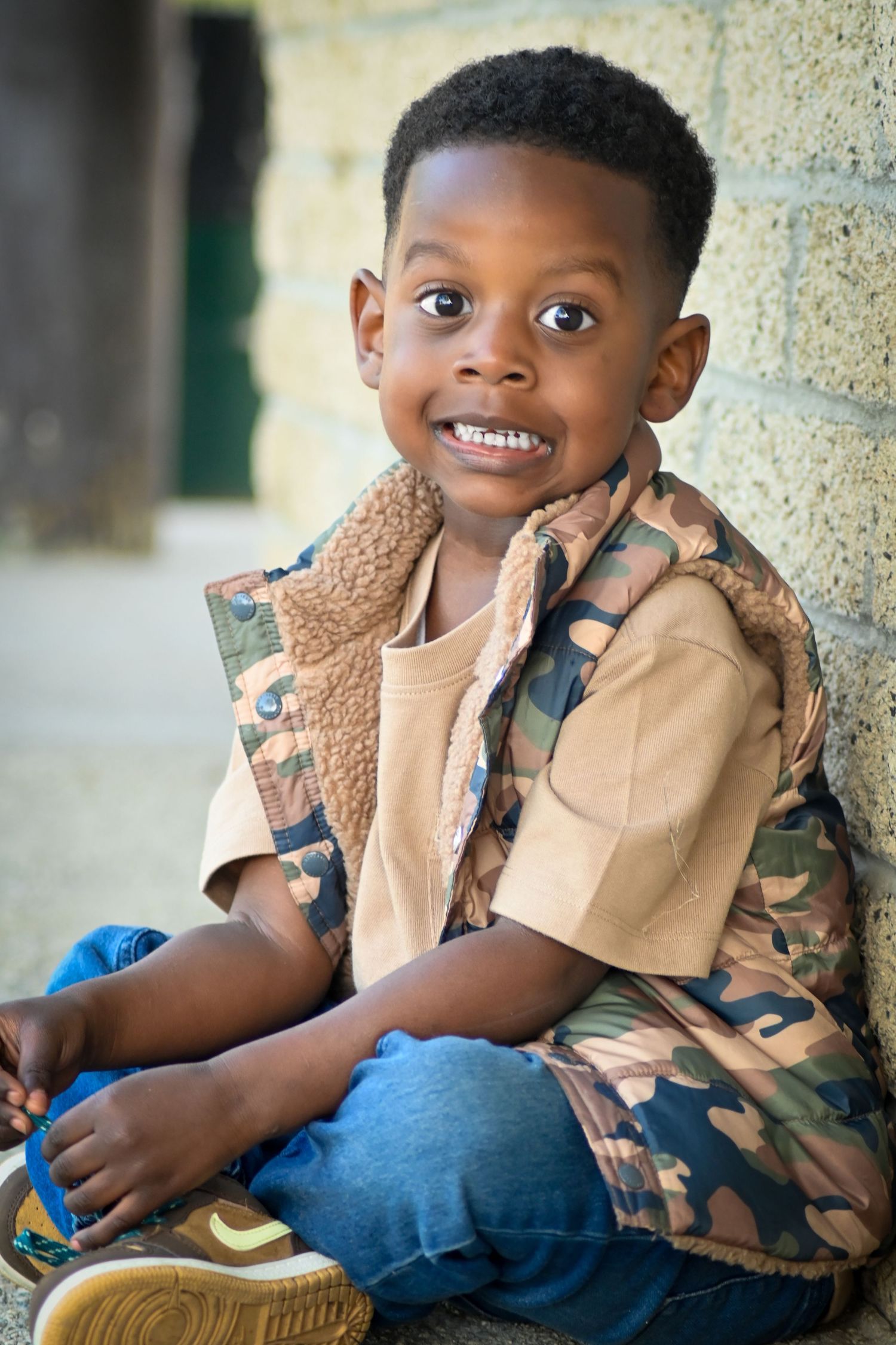 A smiling young Black boy wearing a camouflage vest sits against a stone wall, looking cheerfully at the camera.