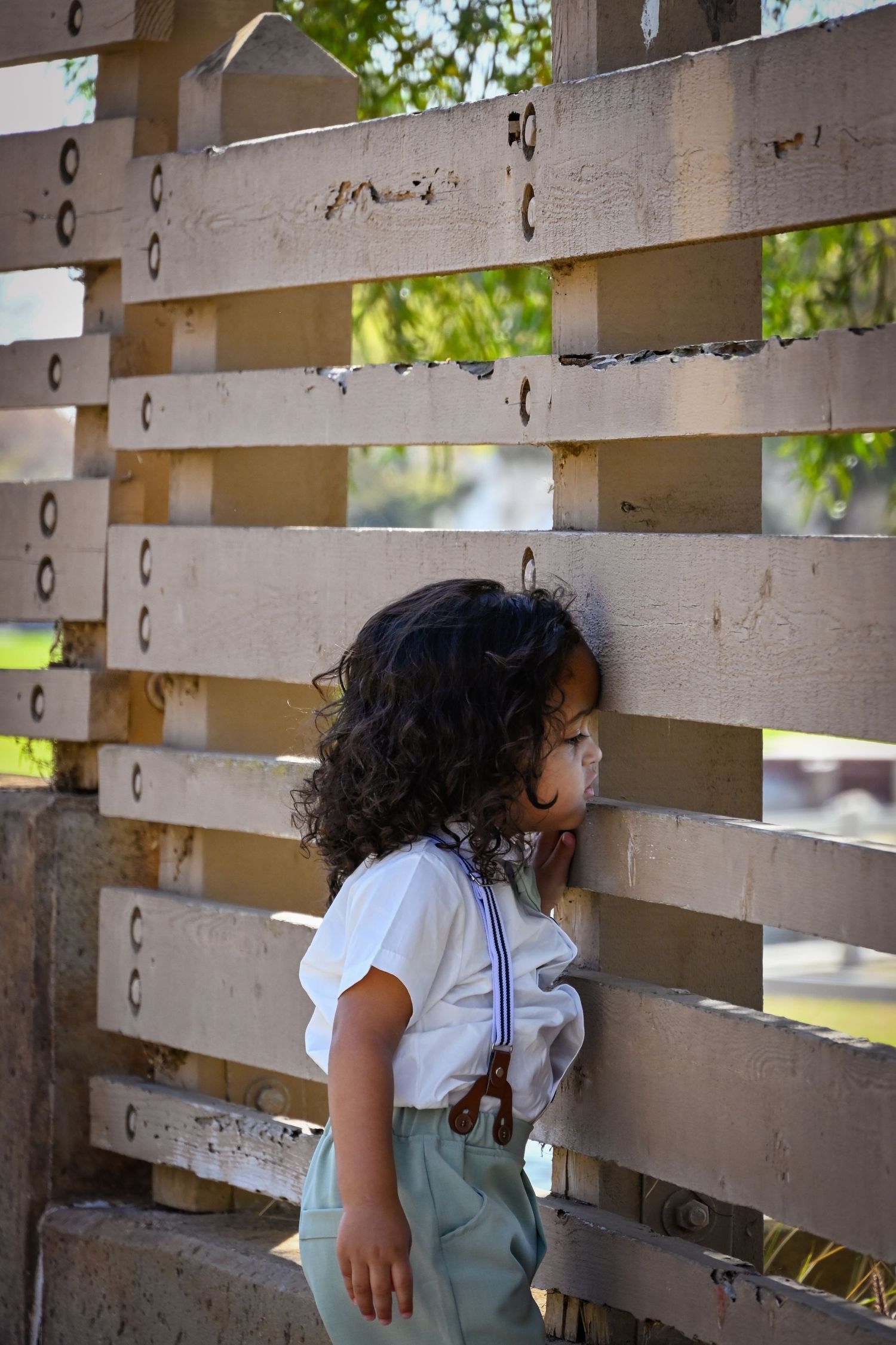 Curly-haired toddler in white shirt and suspenders standing by a wooden slatted fence outdoors.