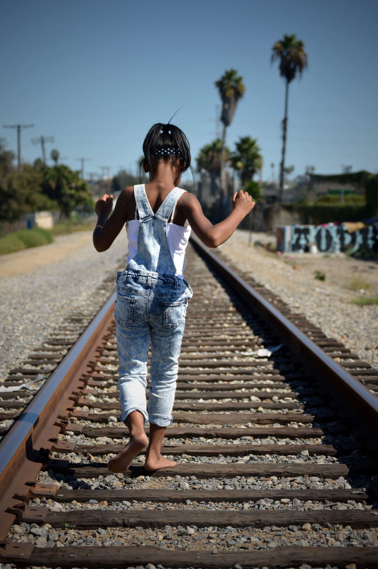 A barefoot child in denim overalls walks along railroad tracks lined with gravel, with palm trees visible in the background.