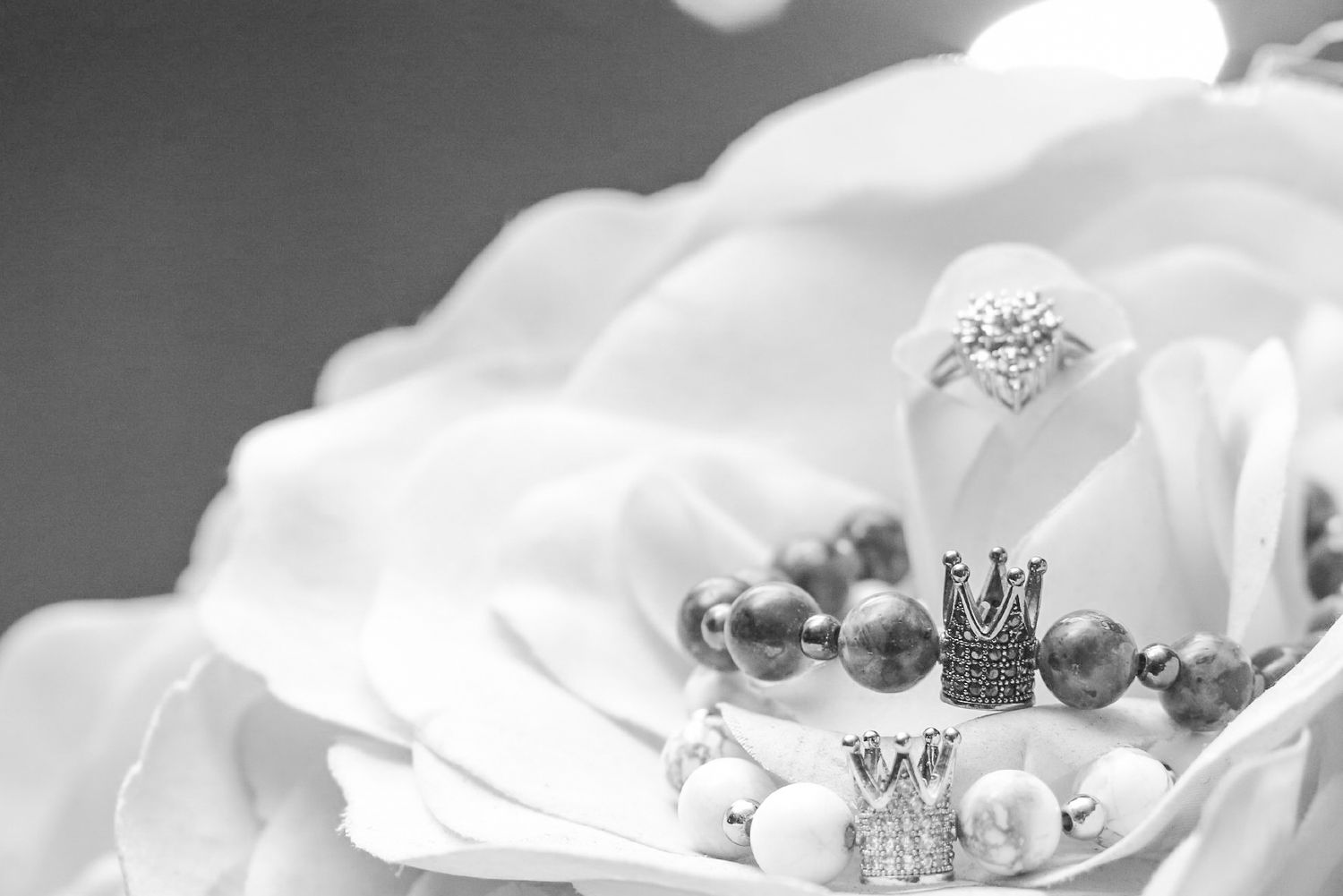 Black and white photo of elegant jewelry including a diamond ring, pearl bracelet, and crown charm on a white rose.