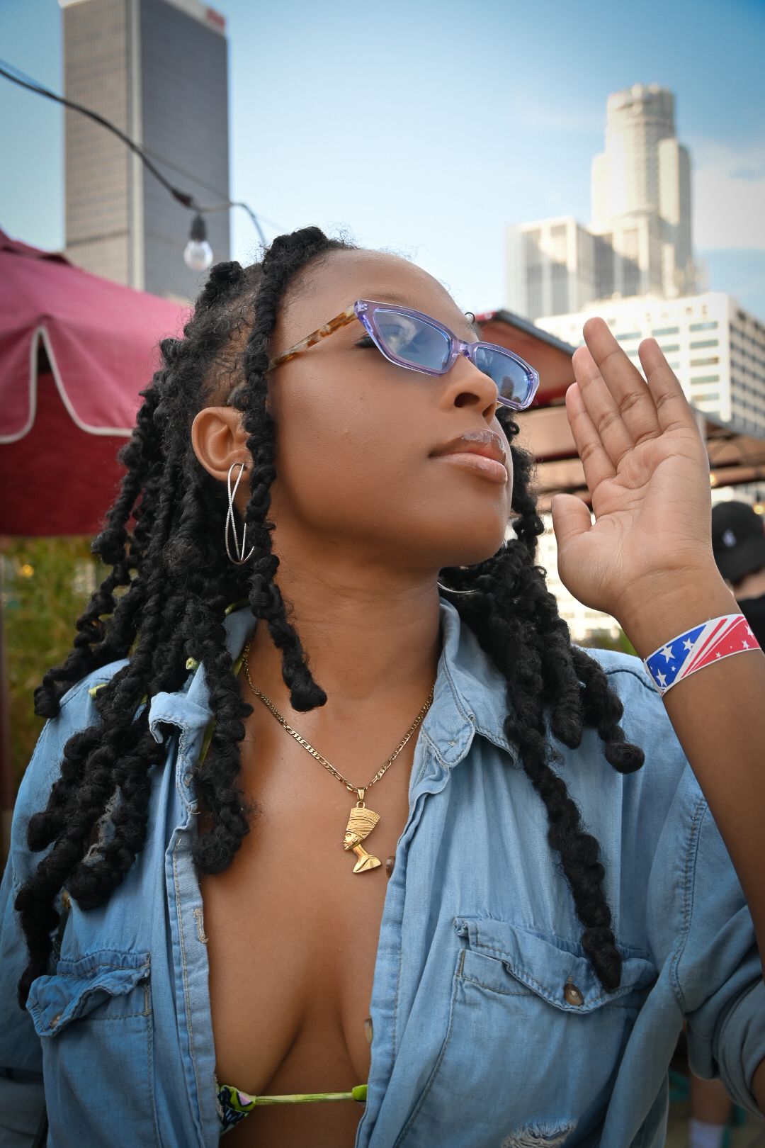 A person wearing sunglasses and denim jacket poses outdoors against an urban backdrop with buildings.