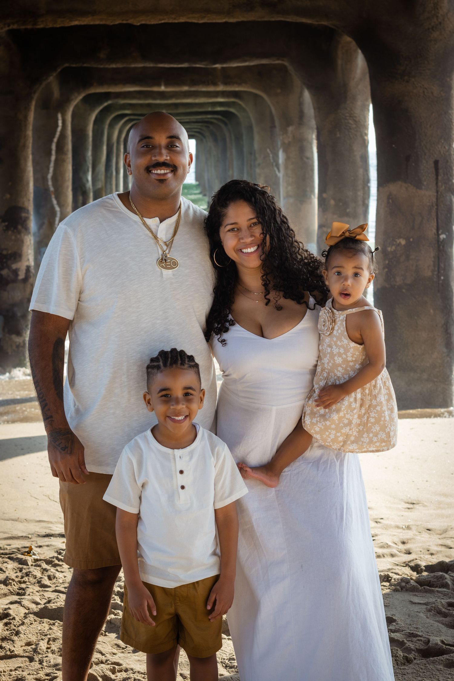 A family wearing coordinated white and beige outfits poses together under a pier at the beach during sunset.