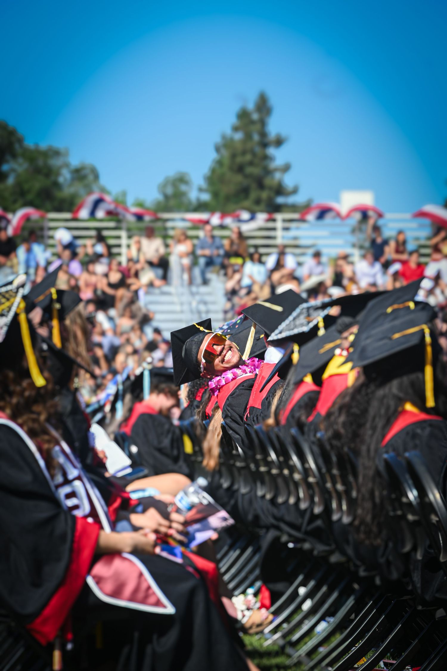 A row of graduates in black caps and gowns sit during an outdoor graduation ceremony under blue skies.