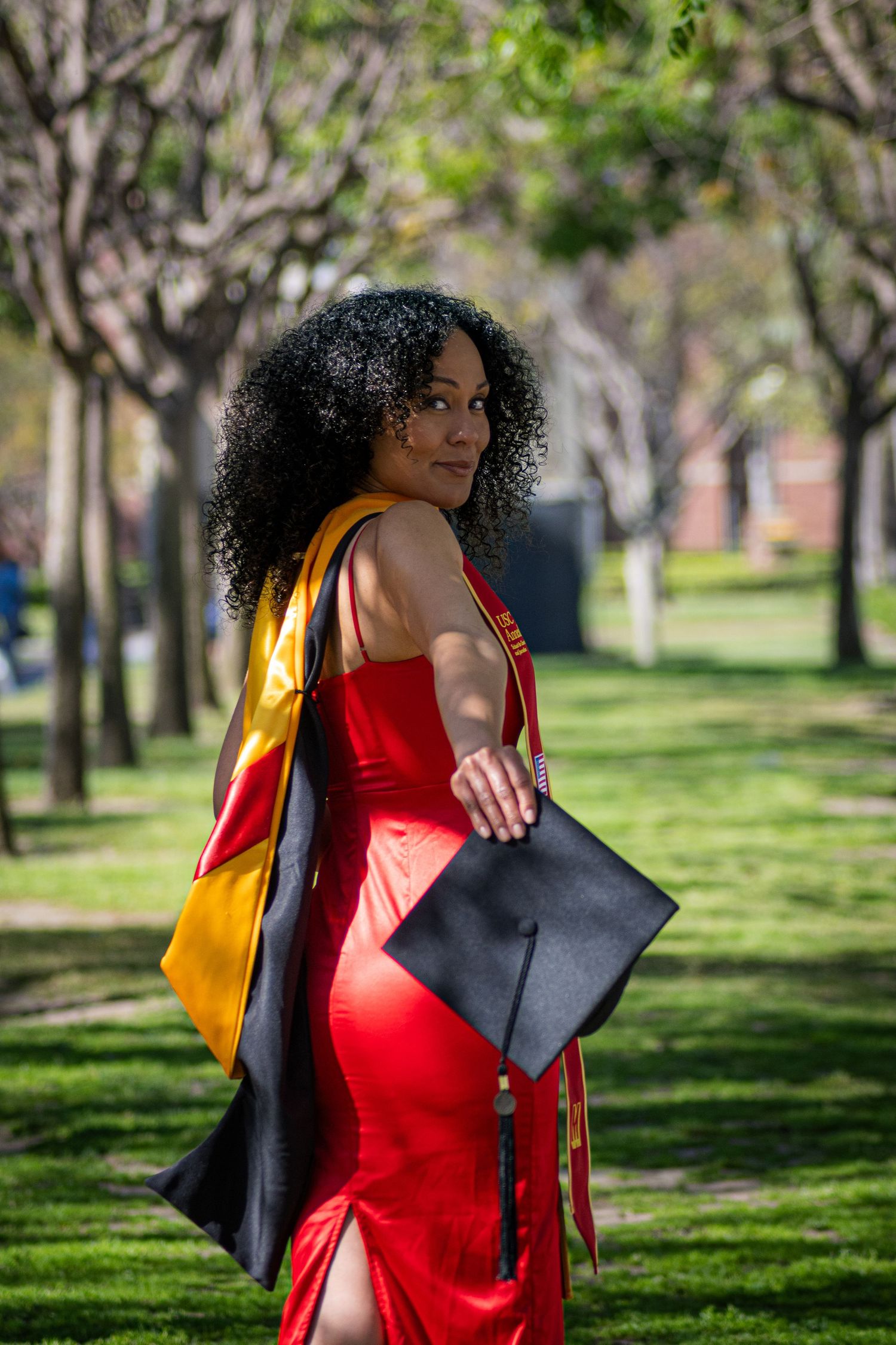Graduate in a red dress walking through a scenic tree-lined campus pathway.