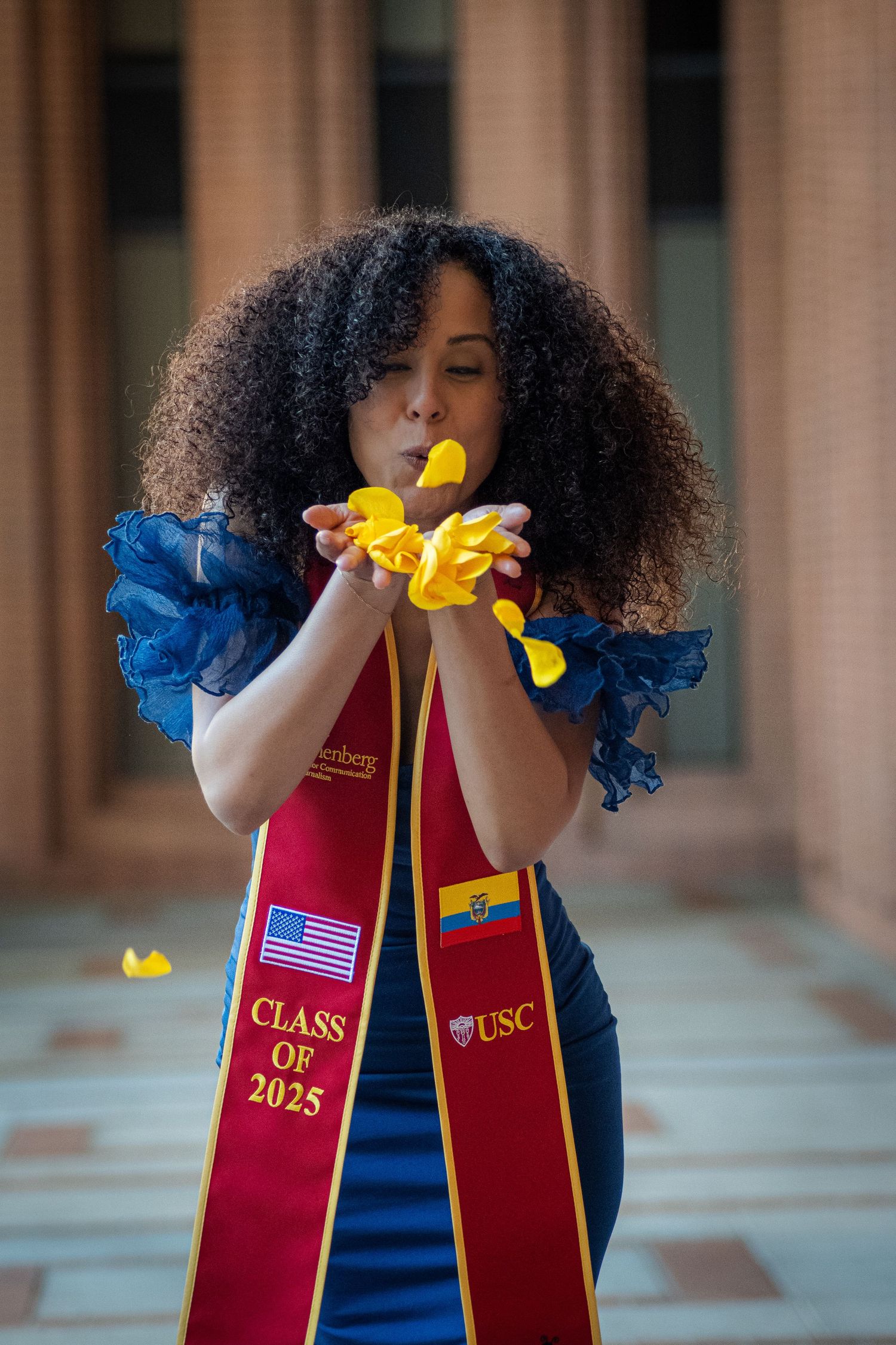 Graduate in blue dress blows yellow confetti while wearing USC Class of 2025 graduation stole.