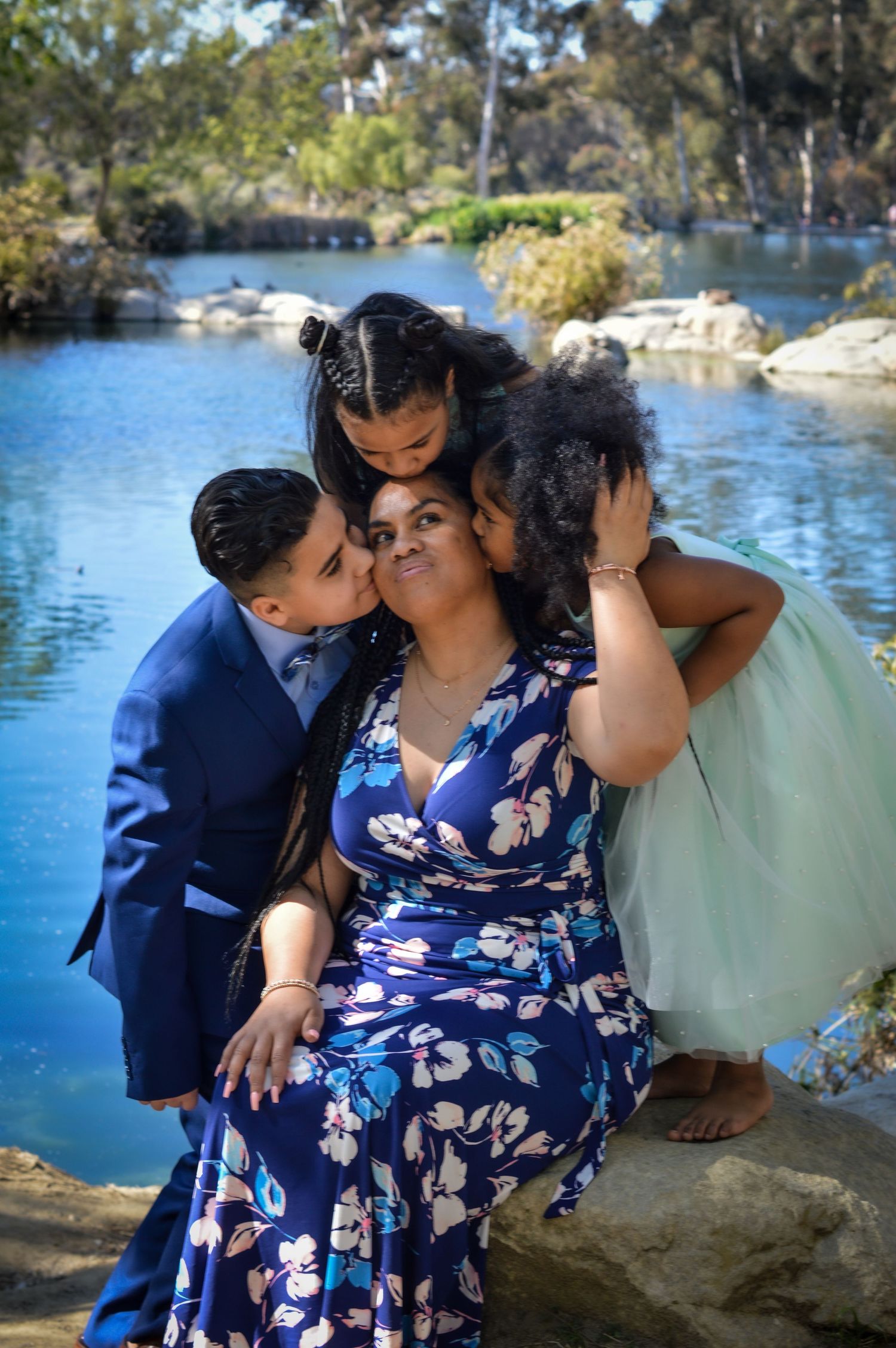 A family in blue and white outfits shares tender moments by a scenic lake on a sunny day.