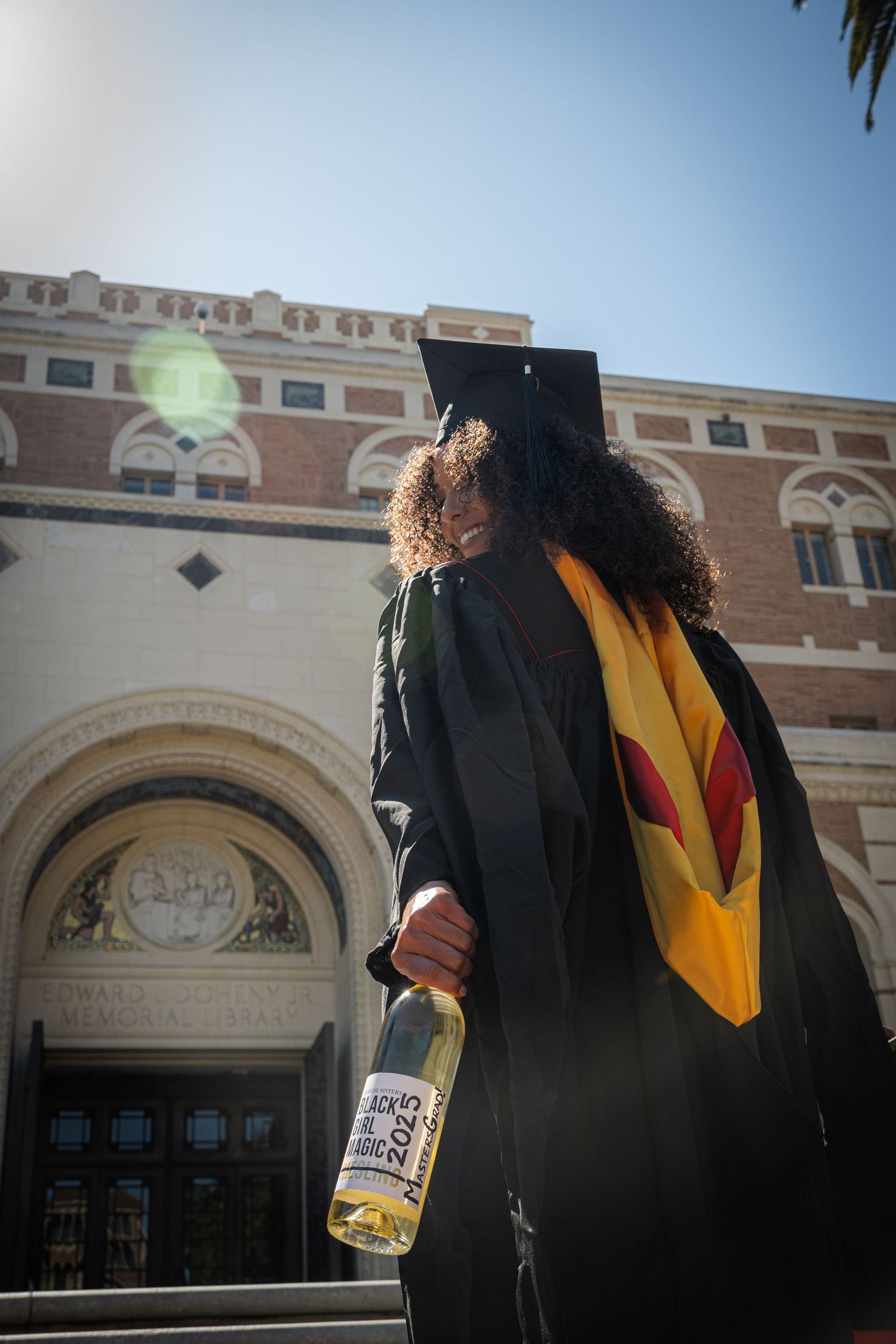 Graduate in cap and gown holding champagne bottle outside university building with classical architecture.