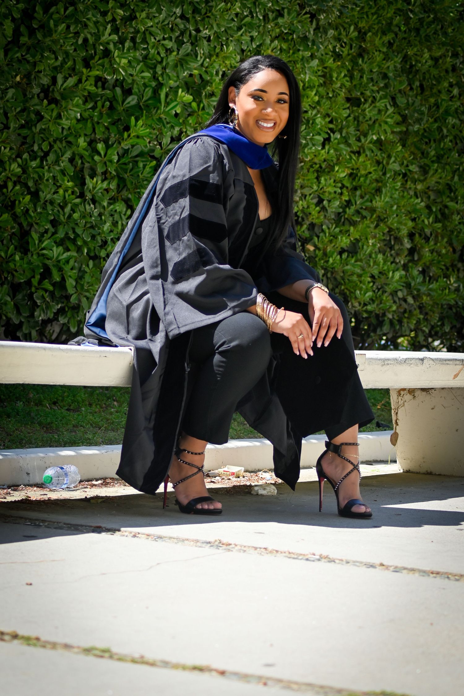 Graduate in black regalia poses while sitting on white ledge outdoors.