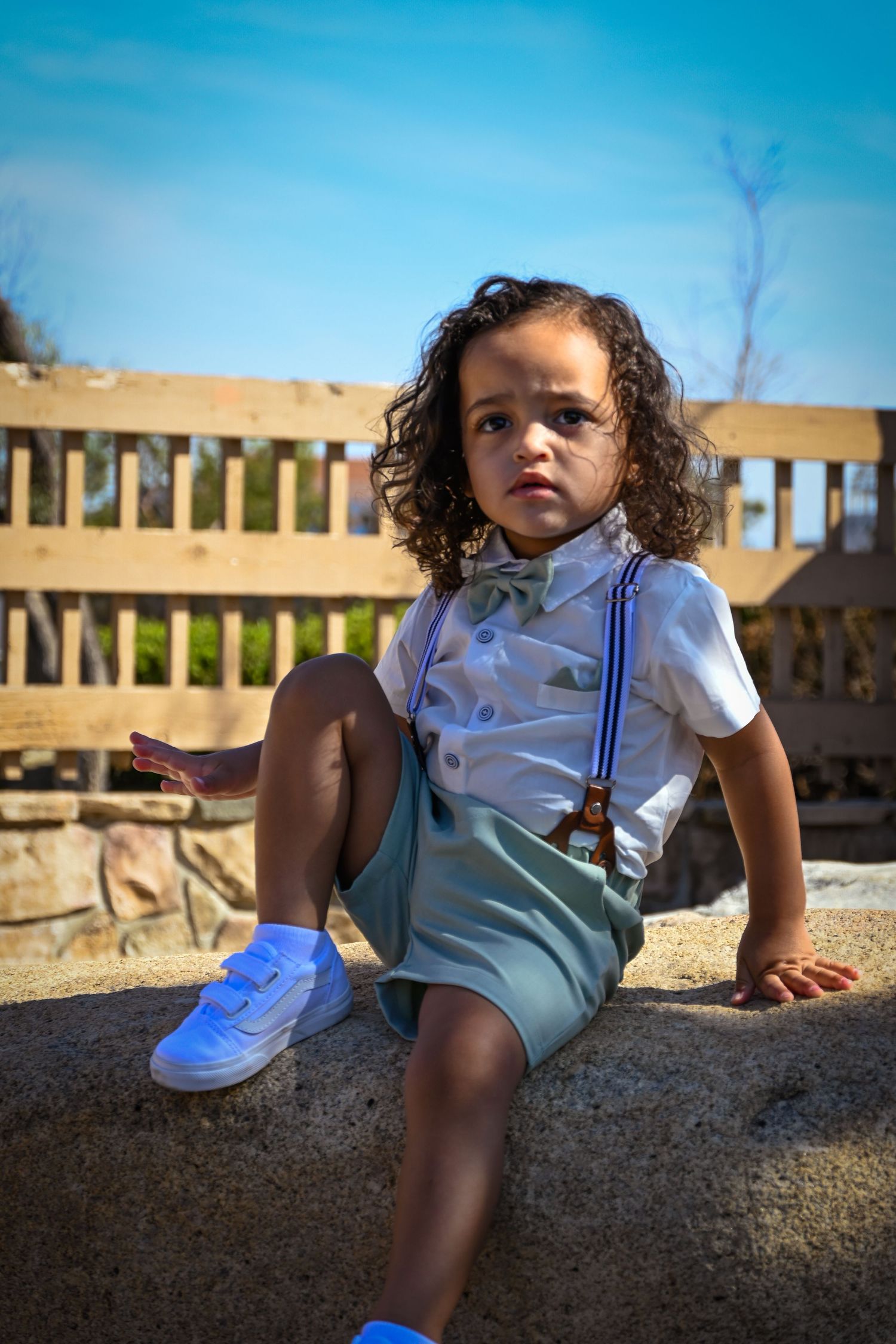 Curly-haired toddler in white shirt, suspenders, and green shorts sitting outdoors on a sunny day.