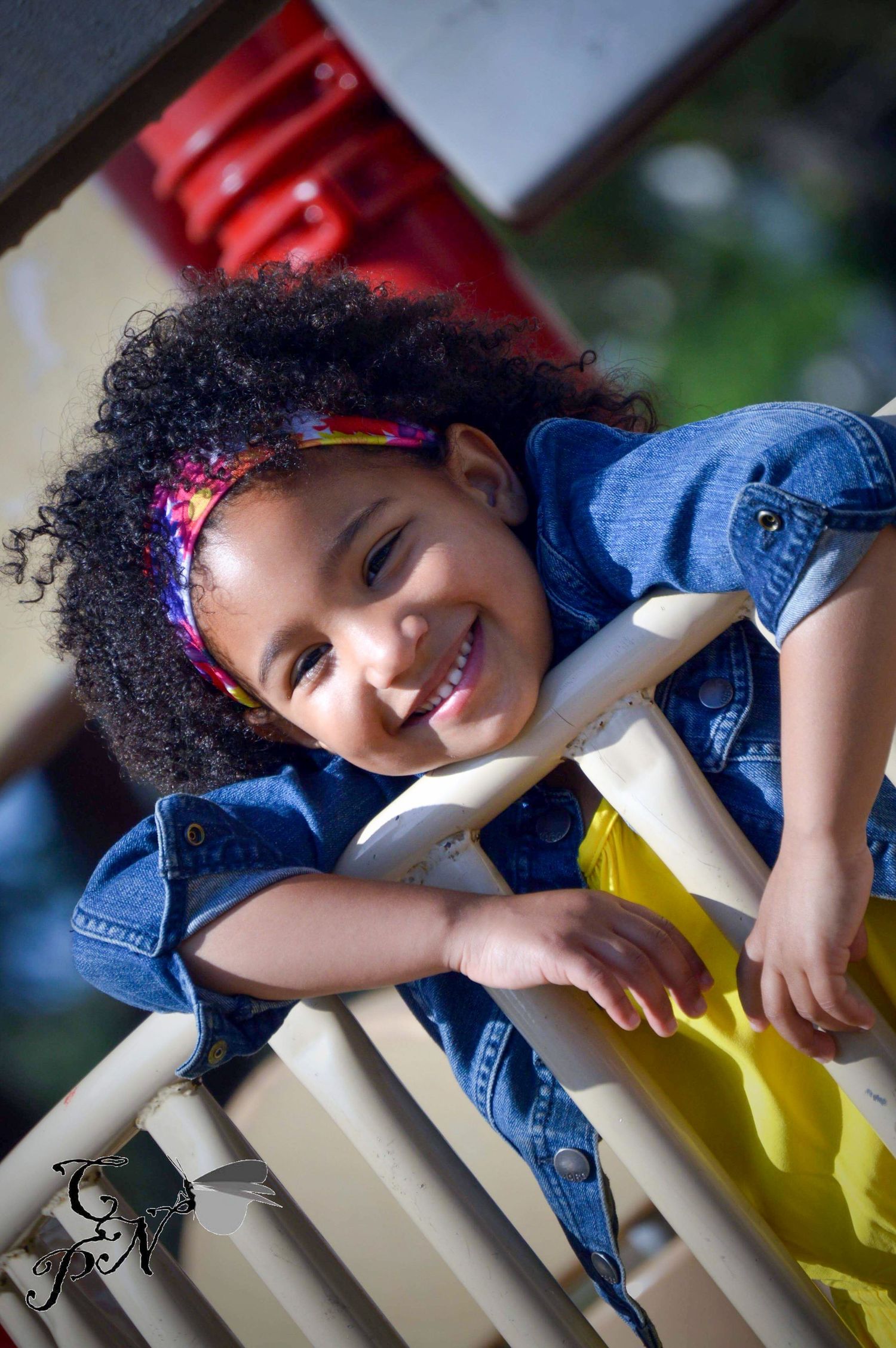 A smiling young girl with curly hair and a colorful headband wearing a denim jacket on a playground.