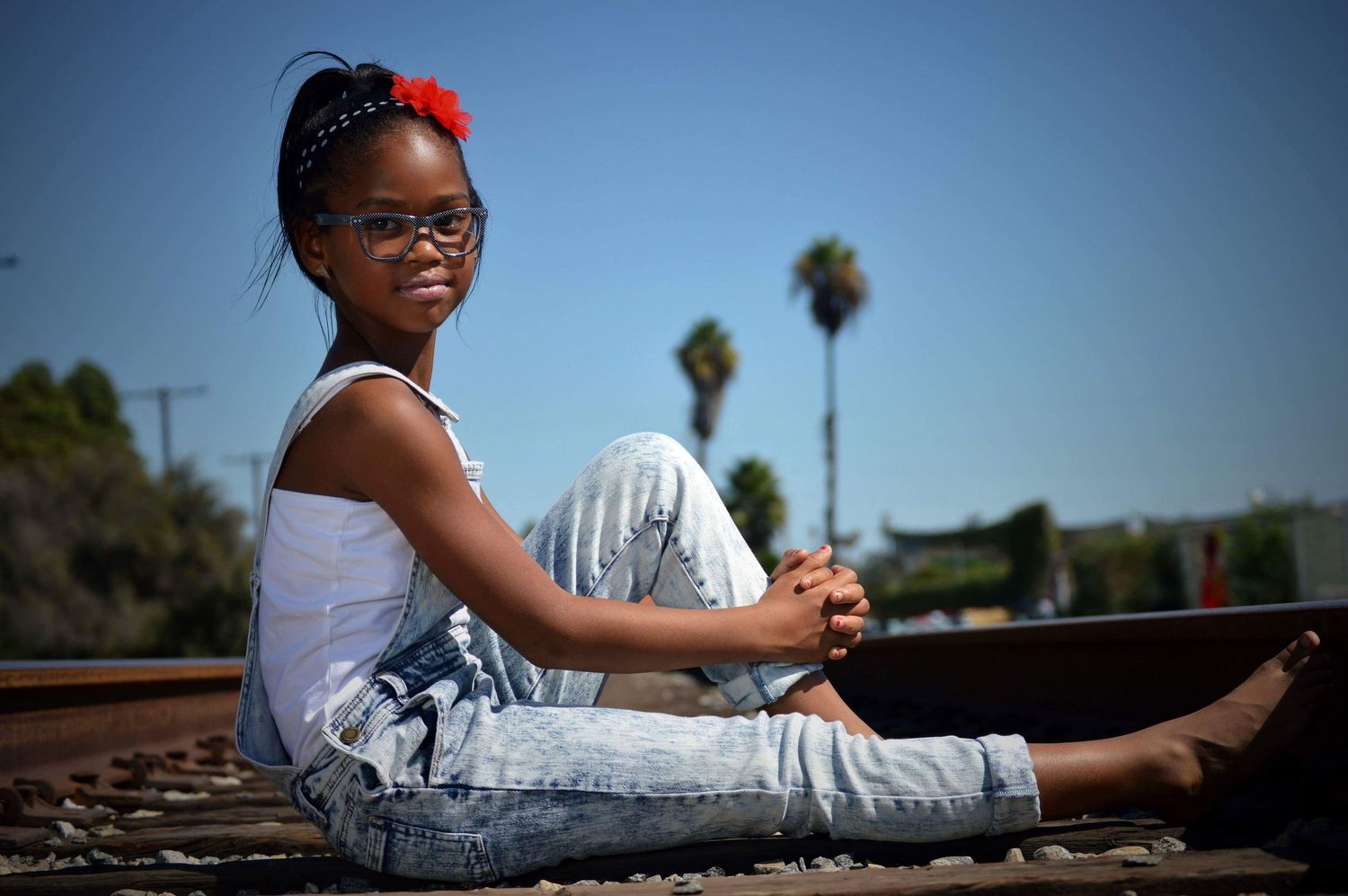 Young girl with glasses and red flower hair clip sits on railroad tracks, smiling confidently on a sunny day.