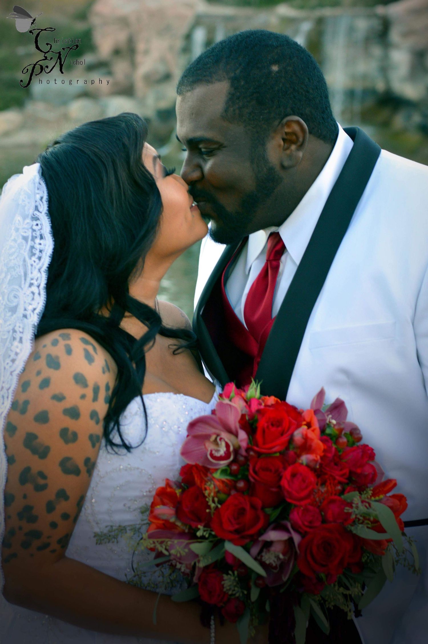 Interracial couple sharing a kiss at their wedding, bride holding red rose bouquet, groom in white suit with red tie.