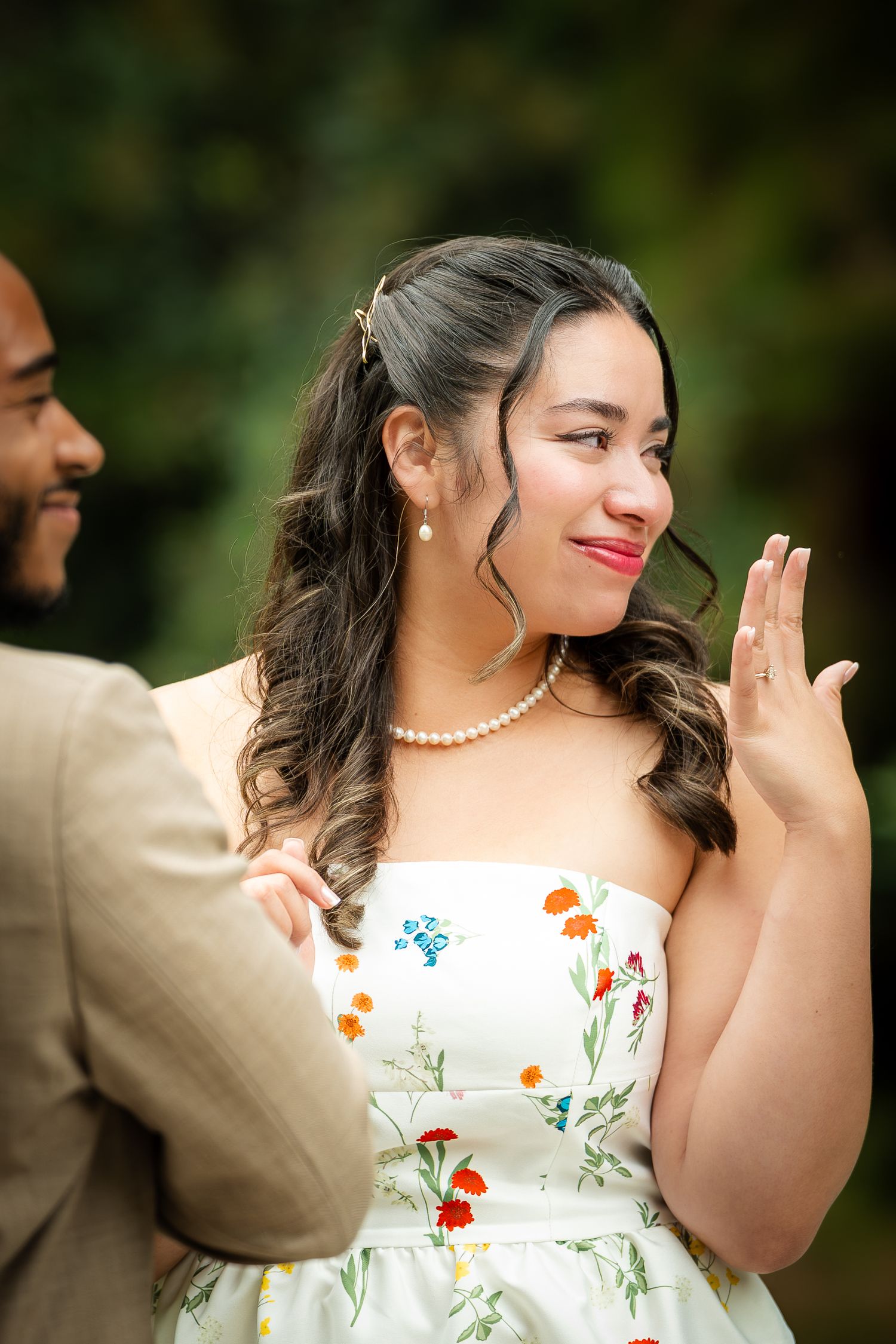 Young woman in floral strapless dress and pearl necklace smiling and showing her hand outdoors.