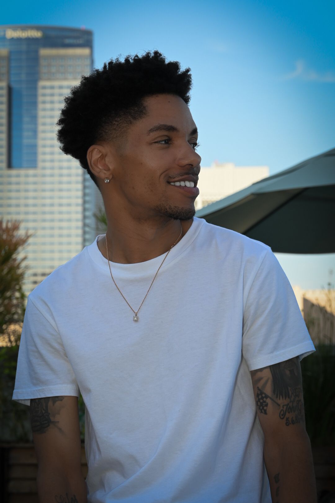A young man in a white t-shirt and necklace smiles while standing outdoors with a city skyline in the background.