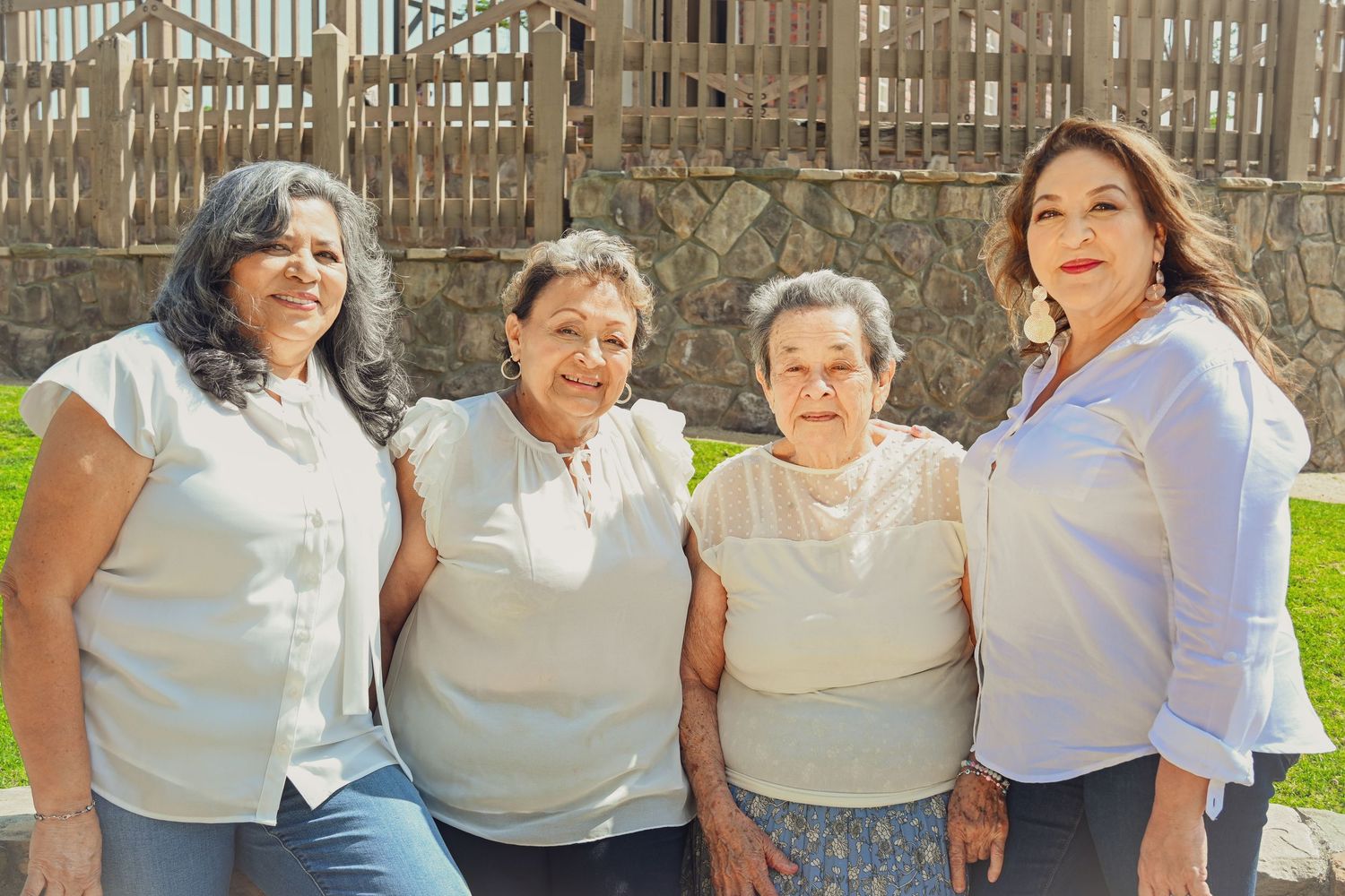 Four people wearing light colored shirts stand together outdoors on a sunny day with a wooden fence in the background.