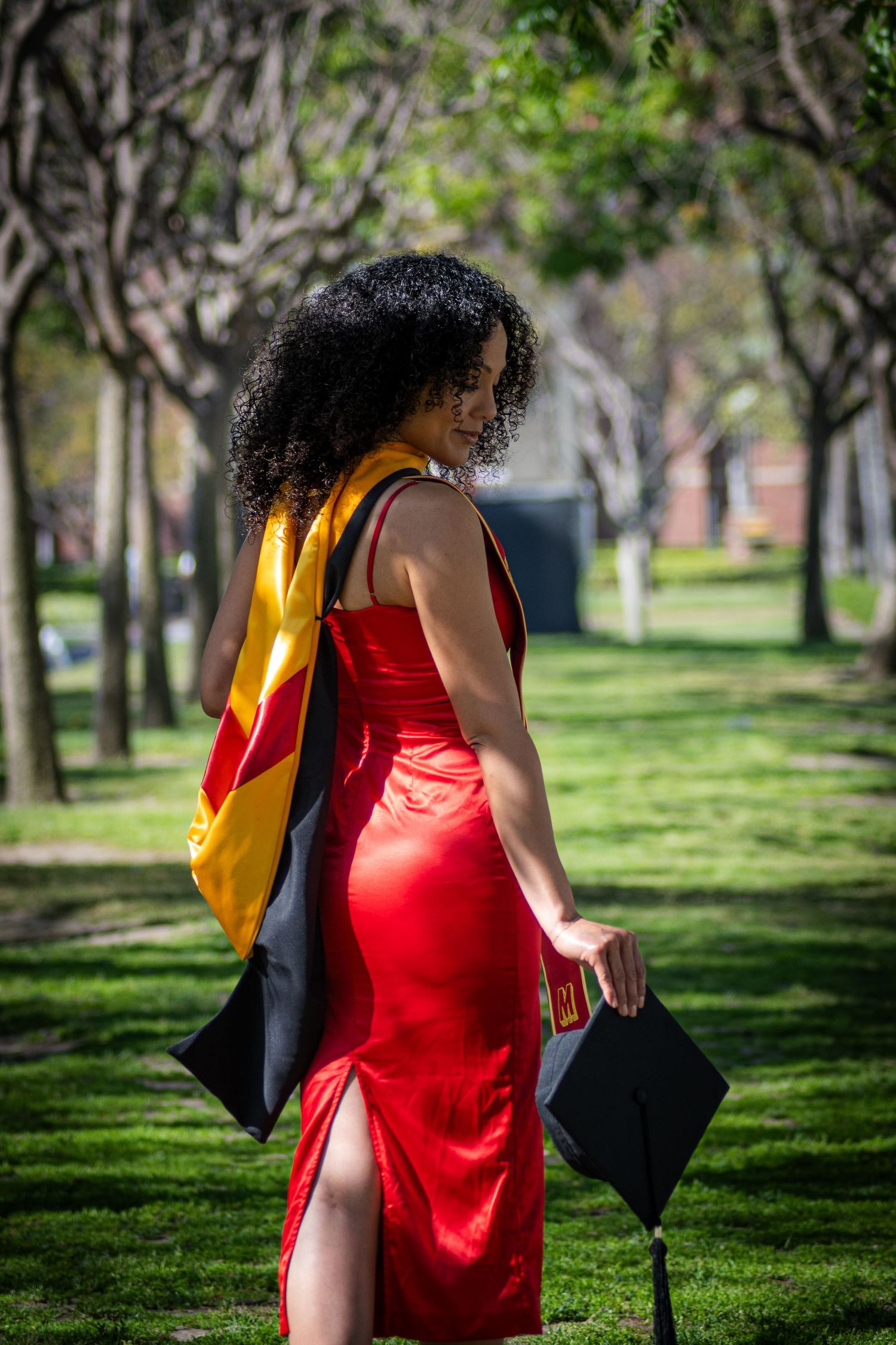 Graduate in red regalia strolling along a green campus path with trees.