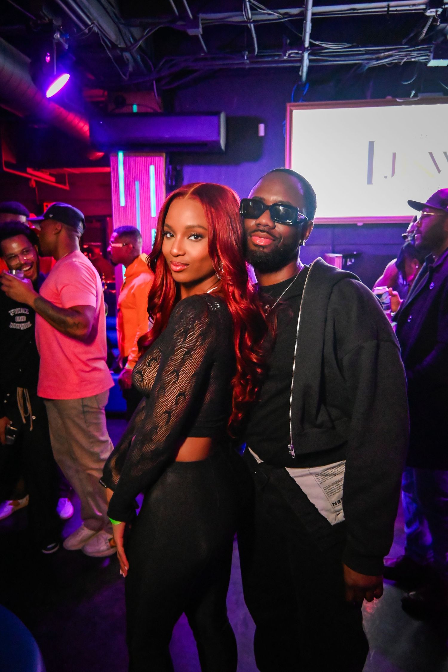 Two people pose together at a nightclub with purple and red lighting in the background.