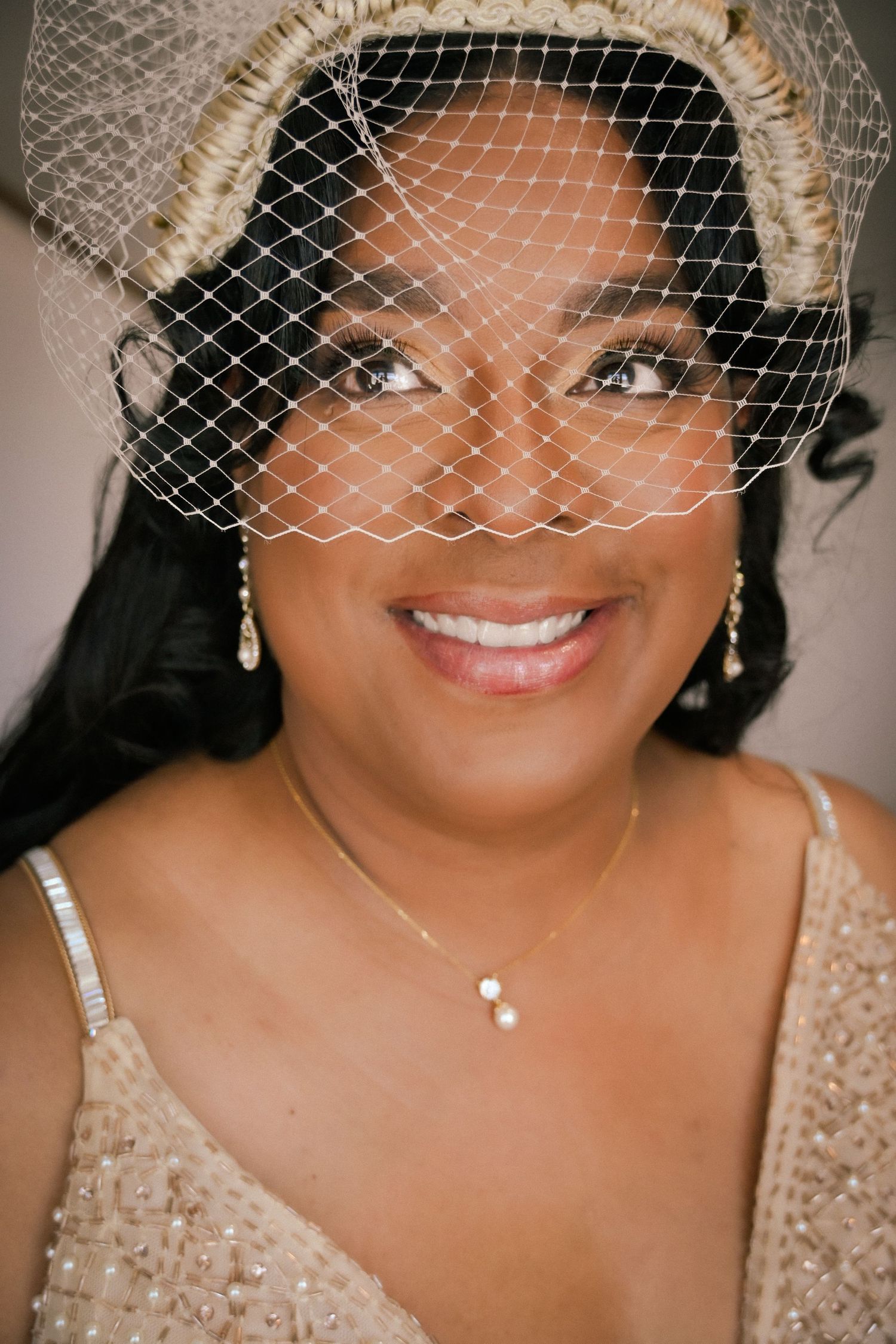Smiling bride wearing a birdcage veil, pearl necklace, and beaded gown, with elegant drop earrings.