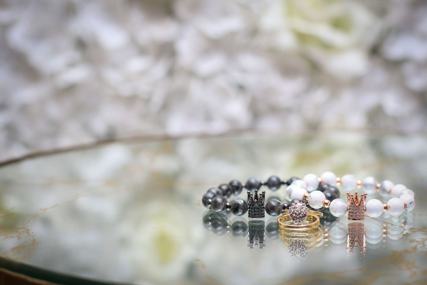 Two elegant beaded bracelets with gold crown charms displayed on a glass surface with white flowers in the background.