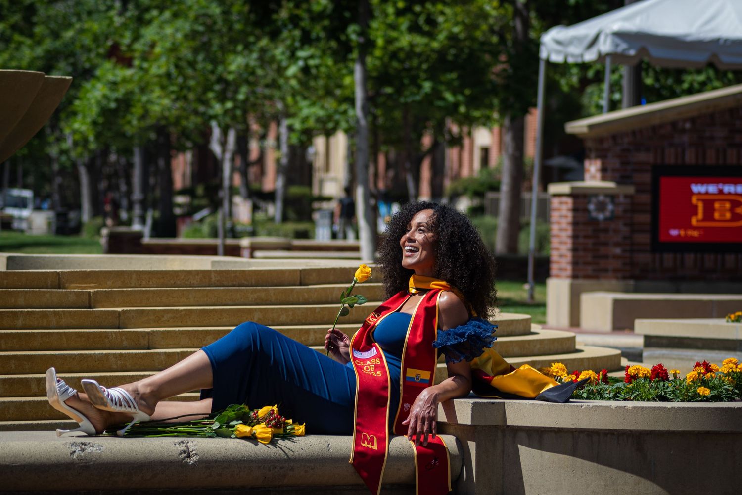 Graduate wearing academic regalia and sash relaxes outdoors on campus grounds.