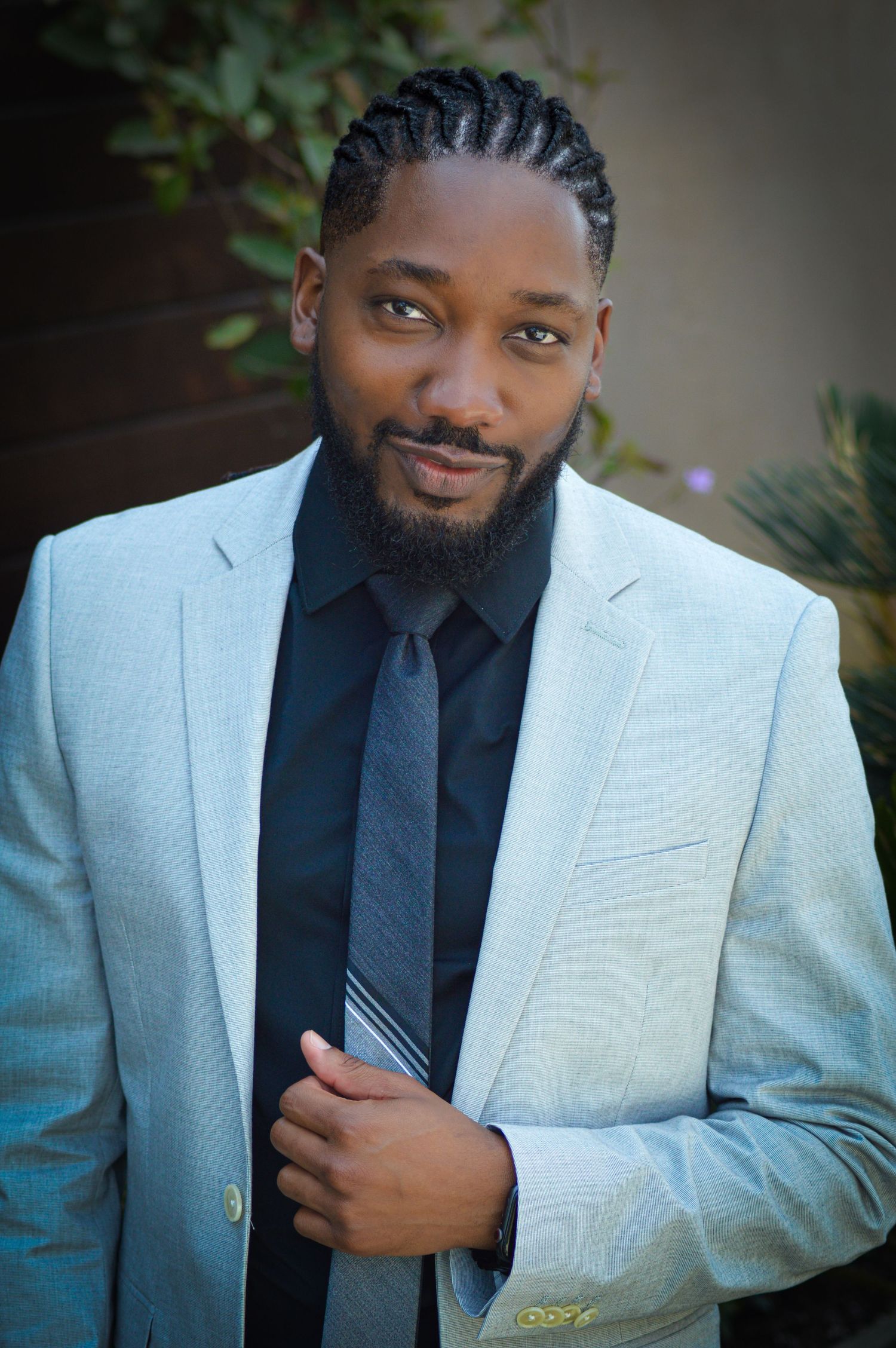 Professional portrait of someone in a light gray suit jacket and dark tie against a blurred outdoor background.