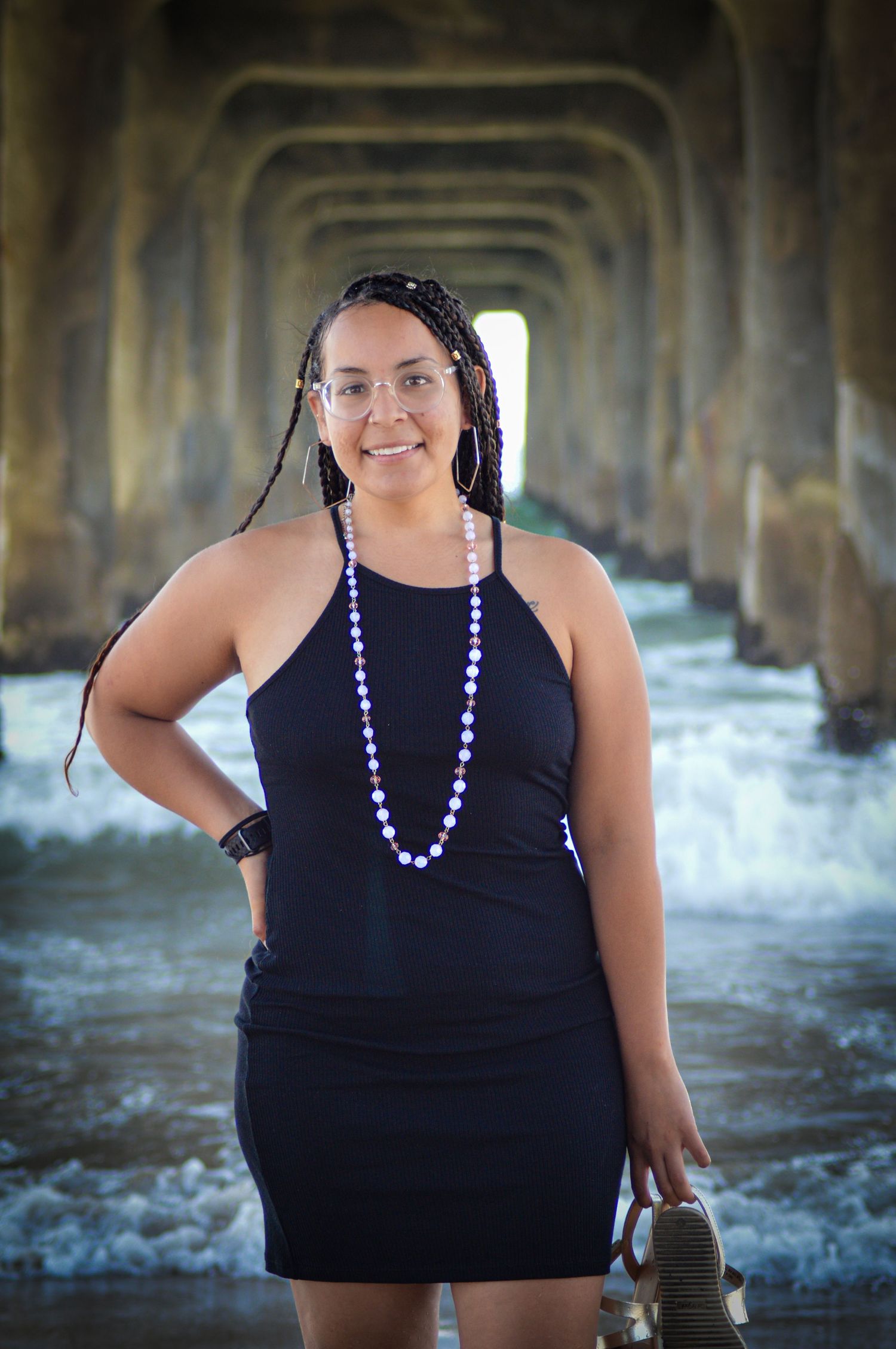 A person wearing a black tank top and purple beaded necklace poses under a pier by the ocean.
