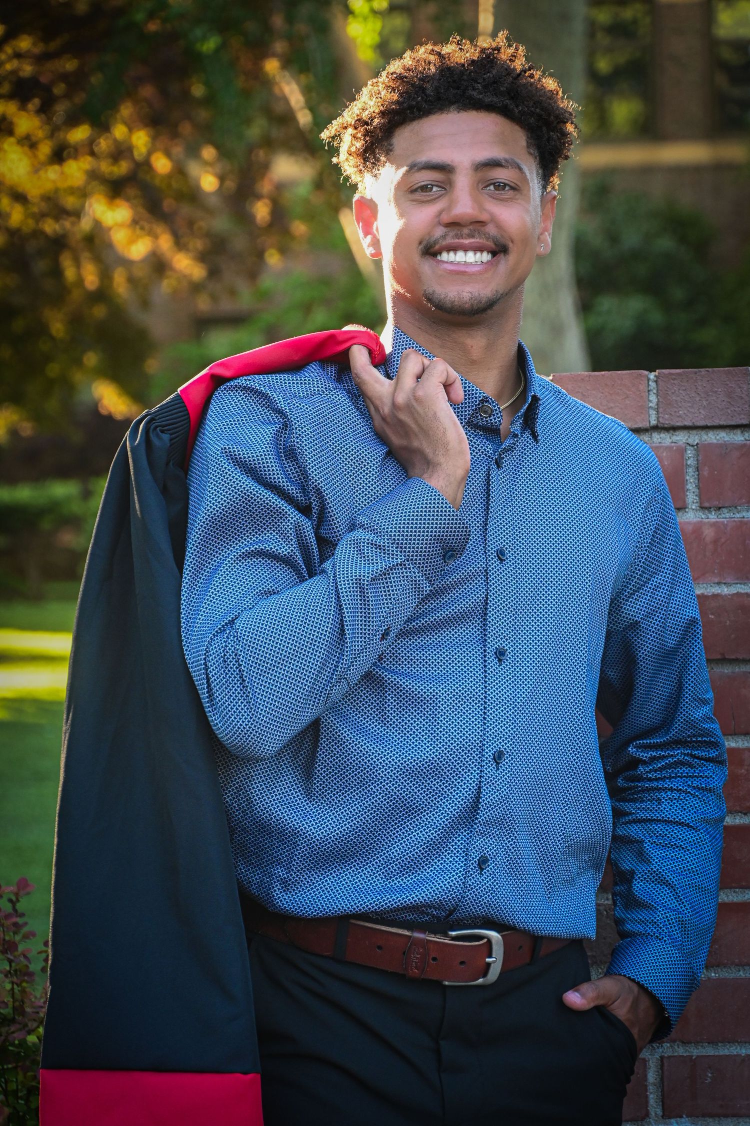 Person in blue button-down shirt and black pants poses outdoors against brick wall during golden hour lighting.