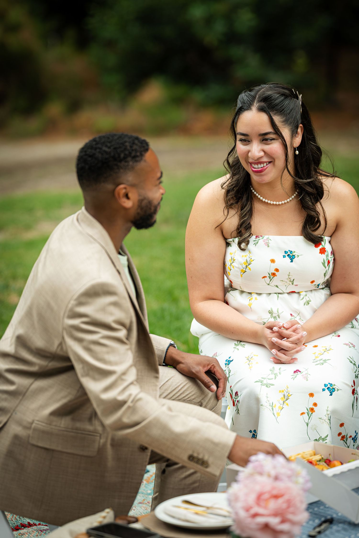 A couple shares a joyful moment at an outdoor picnic, woman in floral dress with pearl necklace, man in tan suit.
