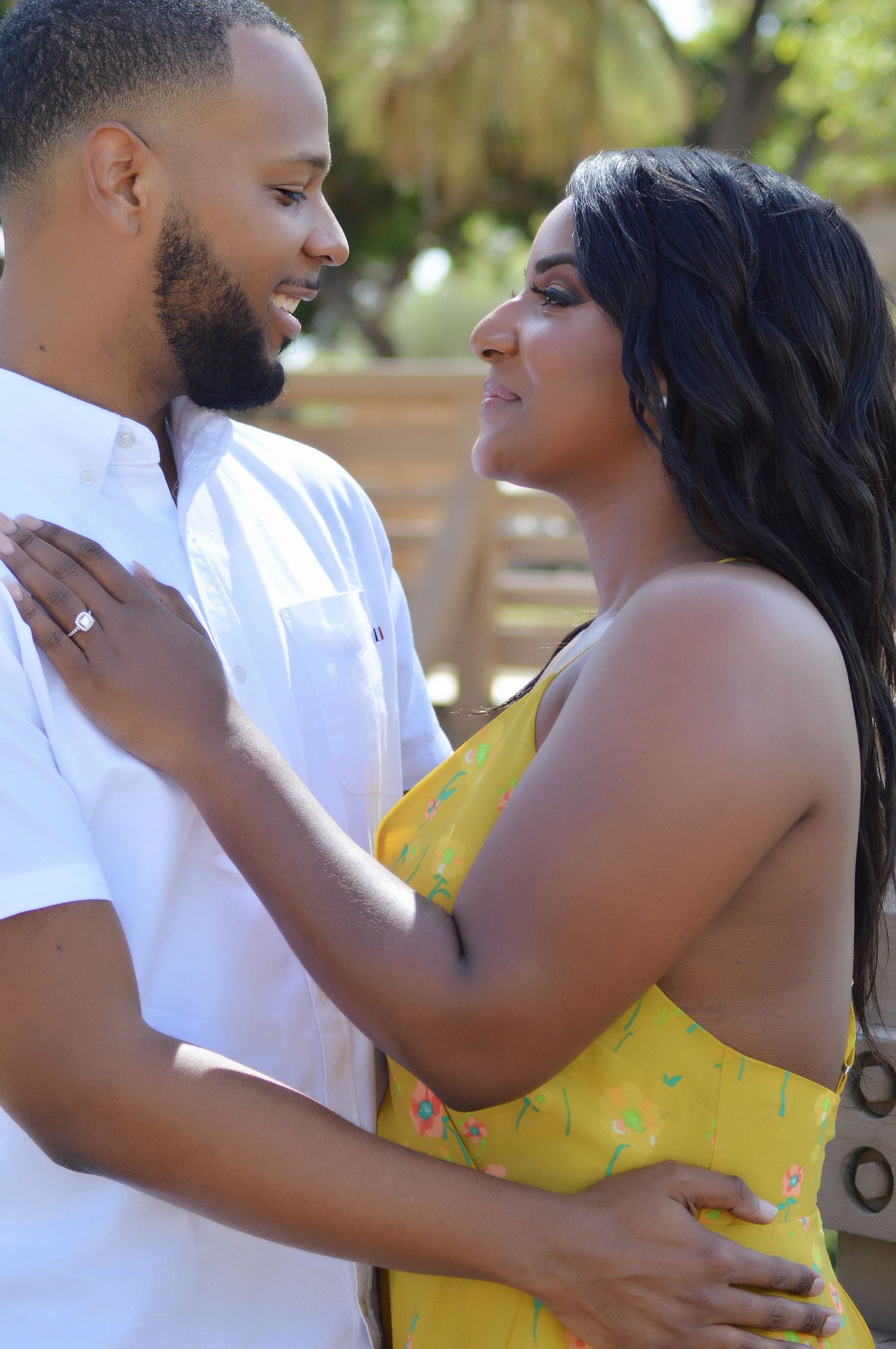 A couple shares a romantic moment outdoors, woman in yellow floral dress and man in white shirt embracing and gazing at each other.