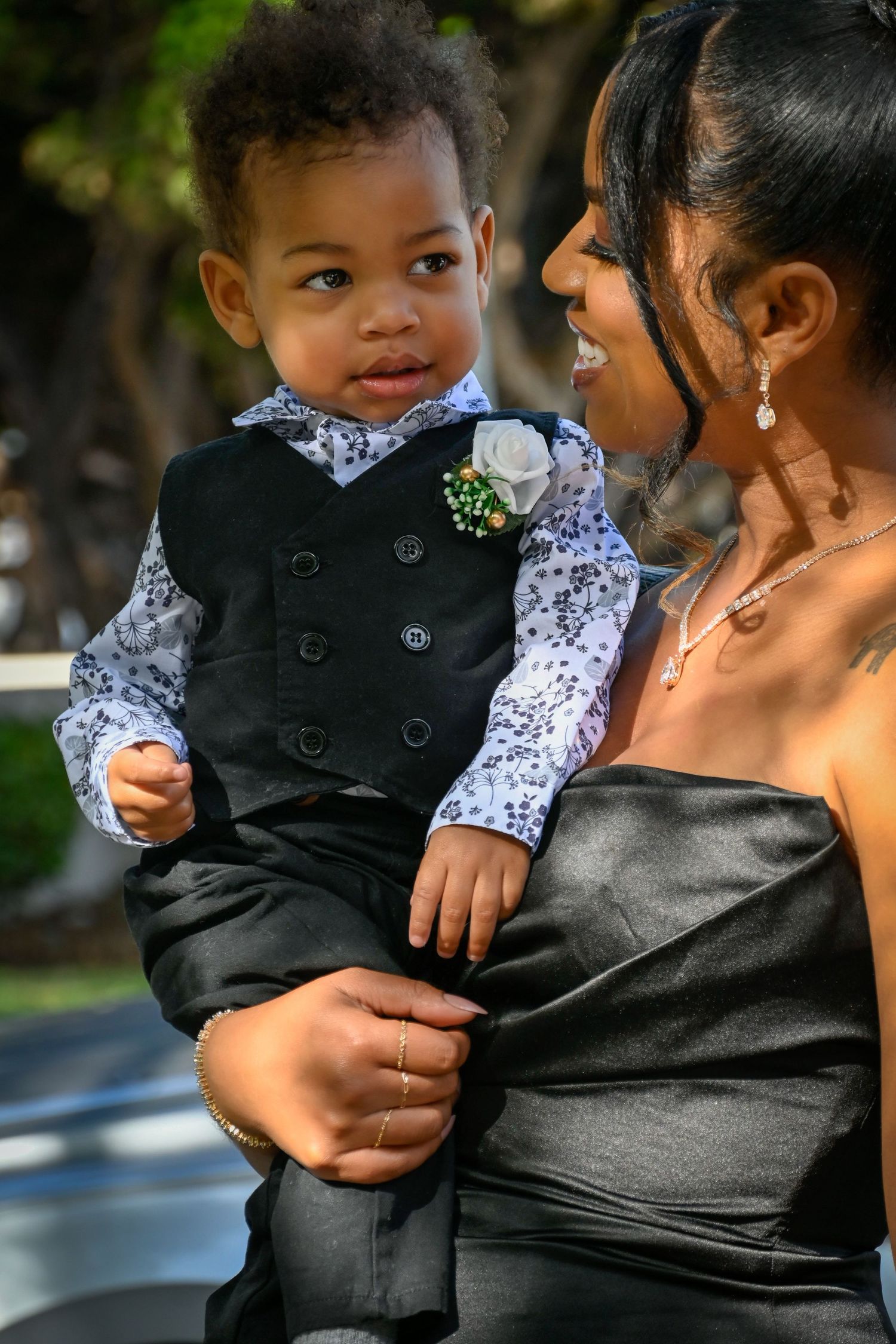 A woman in a black dress holds a well-dressed toddler boy in a black vest and floral shirt with a boutonniere.